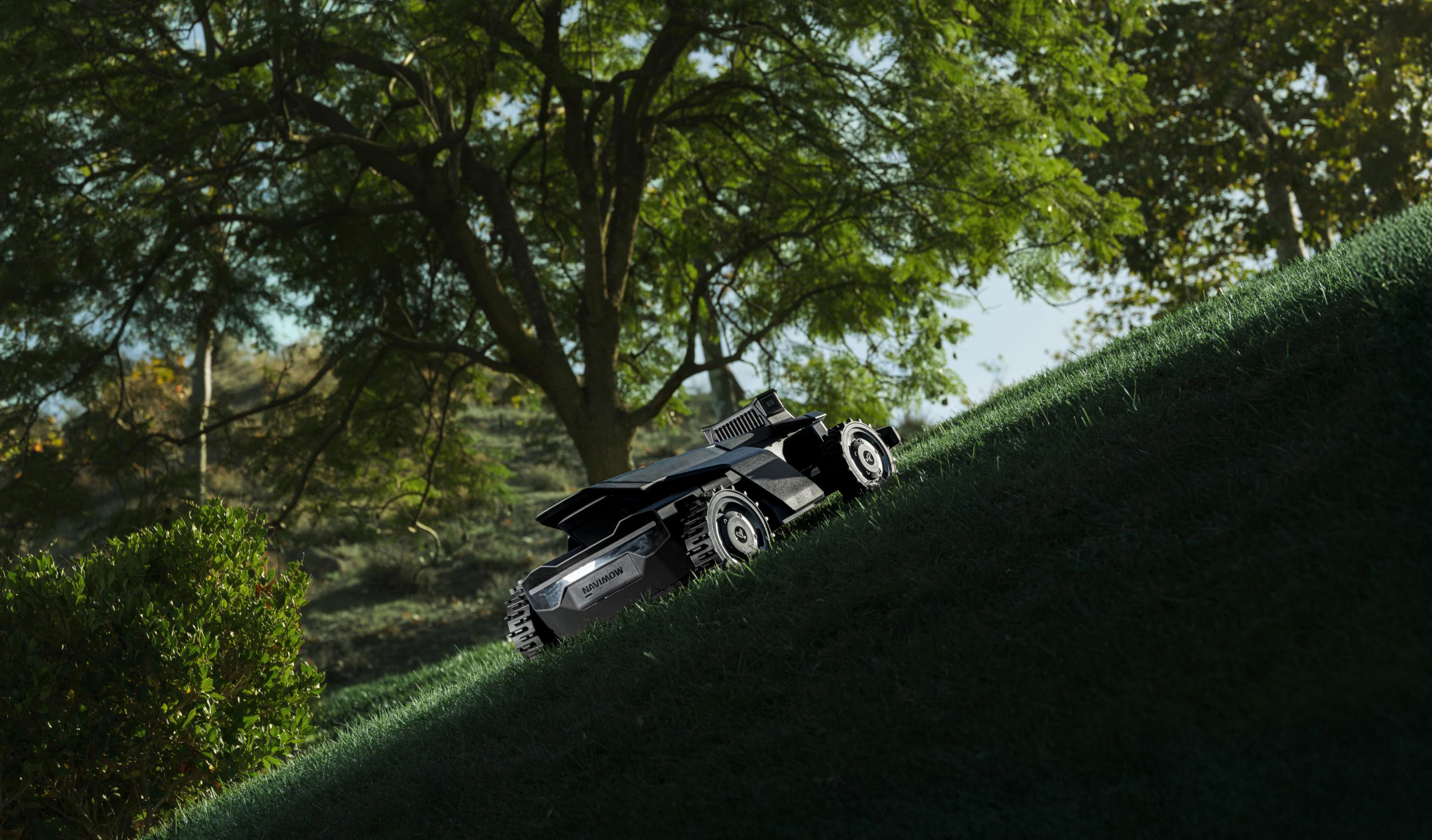 Ein schwarzer Mähroboter auf einem grasbewachsenen Hang mit Bäumen im Hintergrund.