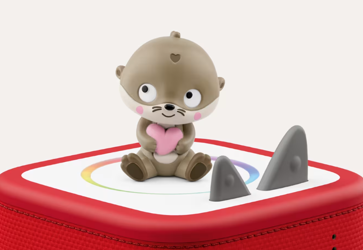 Ein Otter-Spielzeug hält ein rosa Herz auf einem roten Kasten mit Ohren.