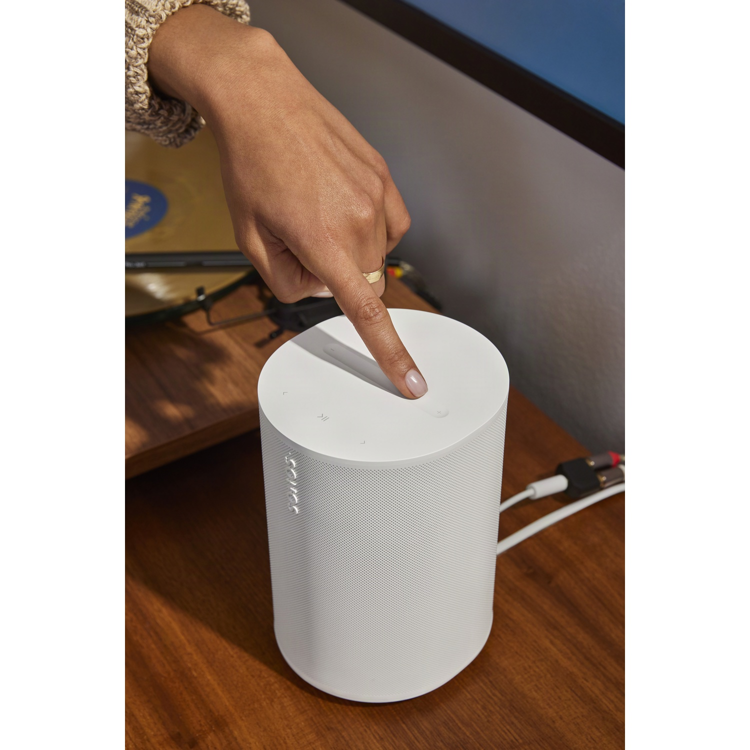 Een witte Sonos speaker op een houten oppervlak, hand drukt op de knop.