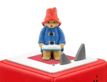Paddington Bär in blauer Jacke mit rotem Hut liest ein Buch, auf dem roten Kasten.