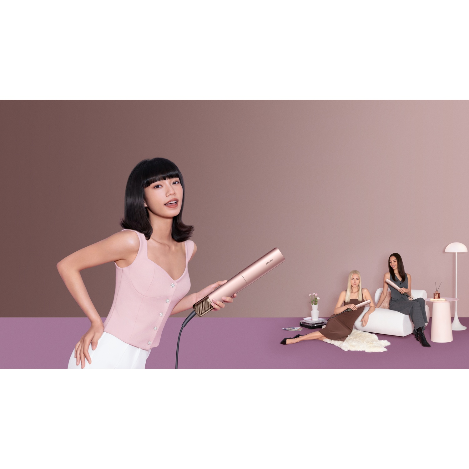 Woman holds pink hair straightener; women sit on sofa in background.