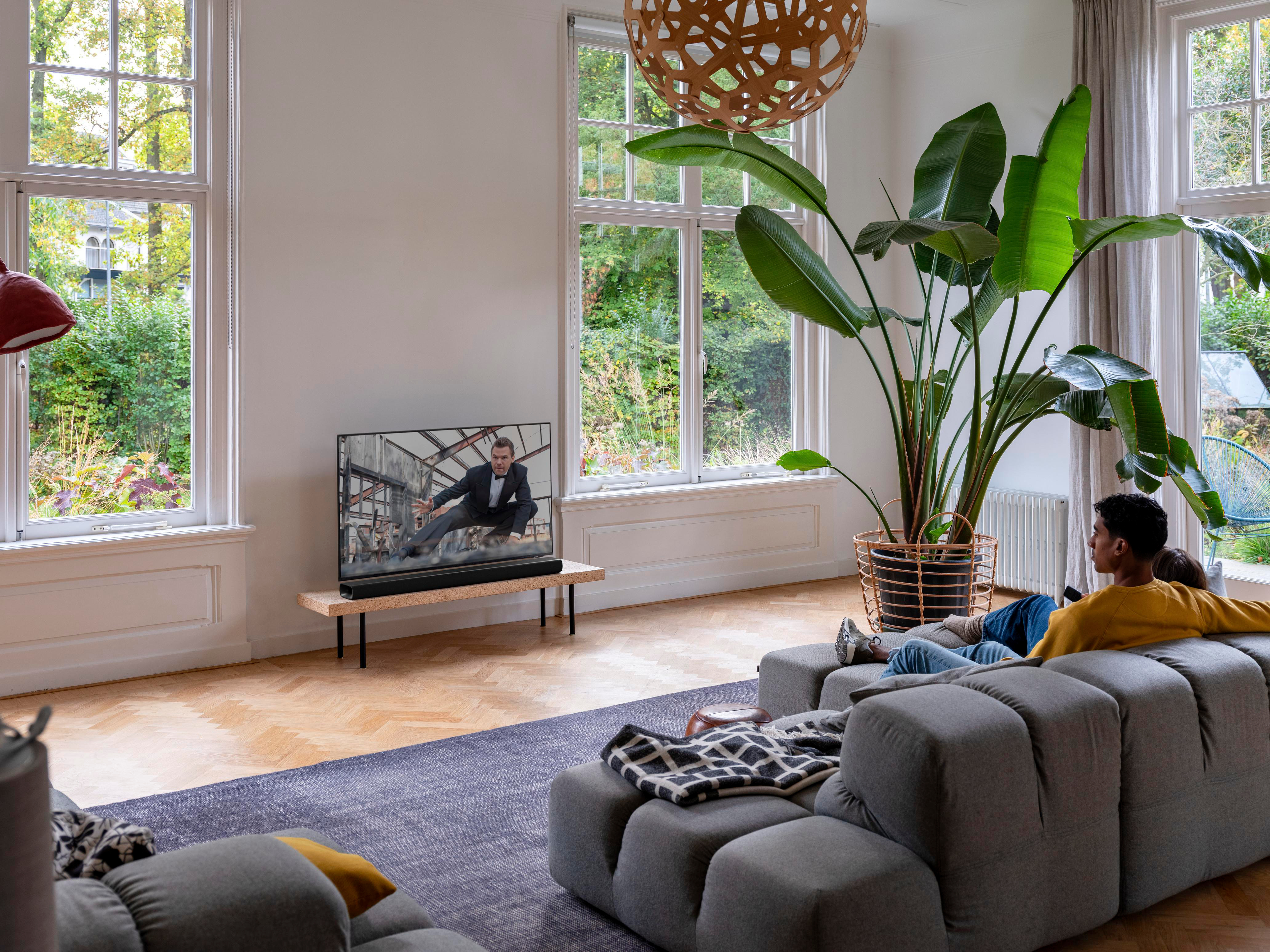 Zwei Personen sitzen auf einem grauen Sofa vor einem Fernseher, der einen Mann im Anzug zeigt.