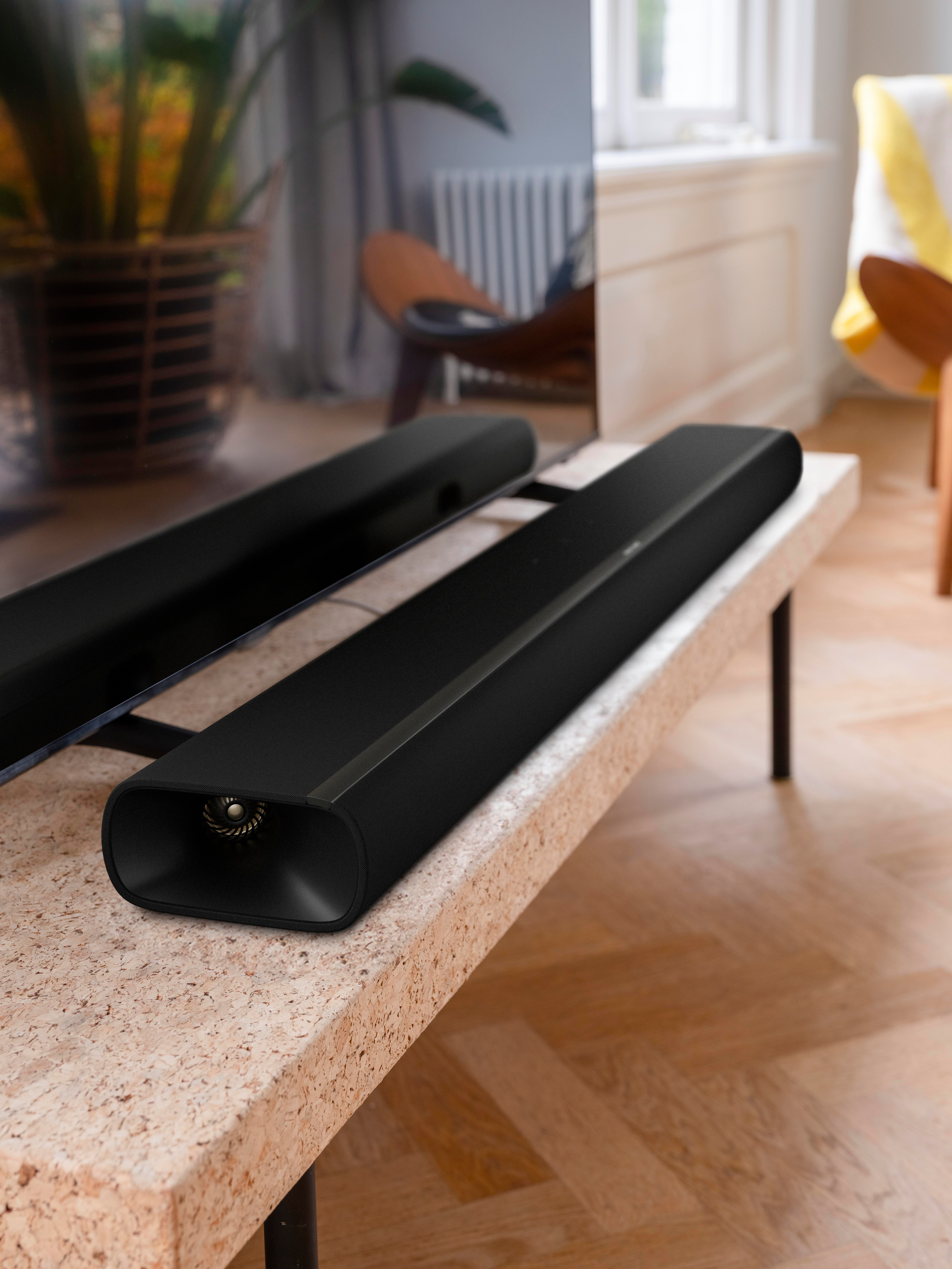 Schwarze Soundbars auf einem Tisch vor einem Fernseher und einem Fenster.