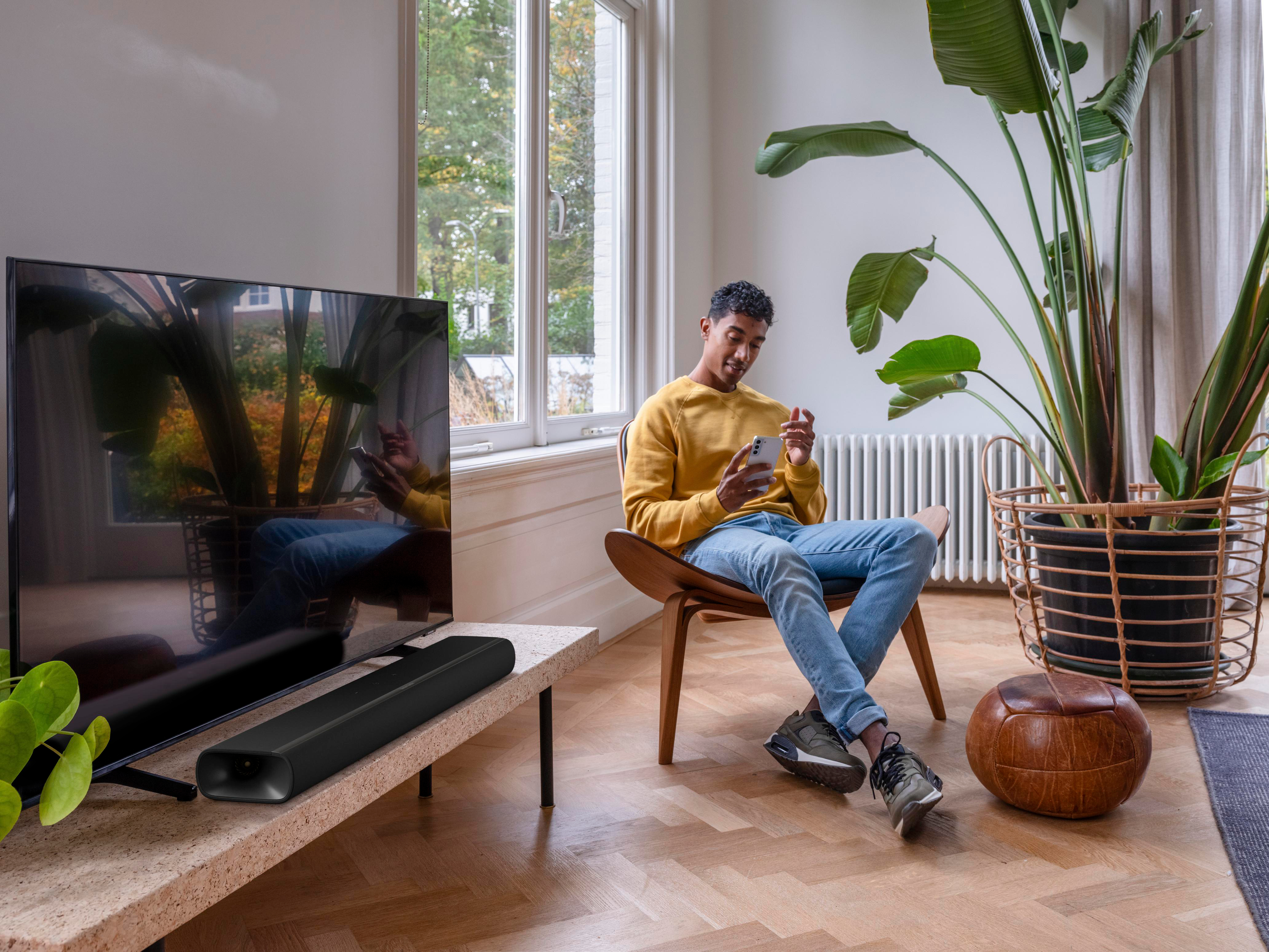 Mann in gelbem Pullover sitzt auf Stuhl und schaut aufs Handy. Fernseher daneben.