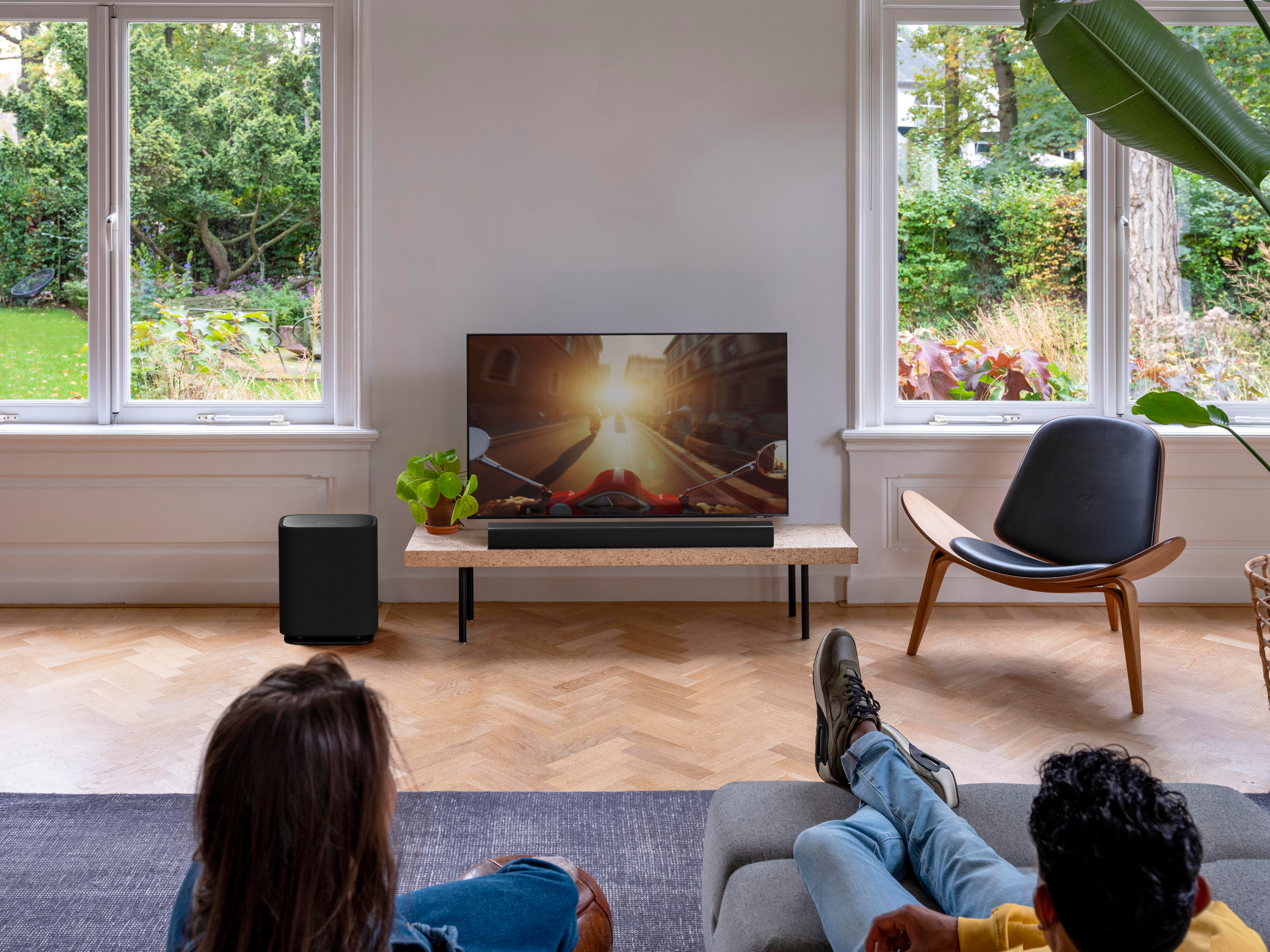 Zwei Personen sitzen auf Sofas und schauen fern. Ein schwarzer Subwoofer steht links.