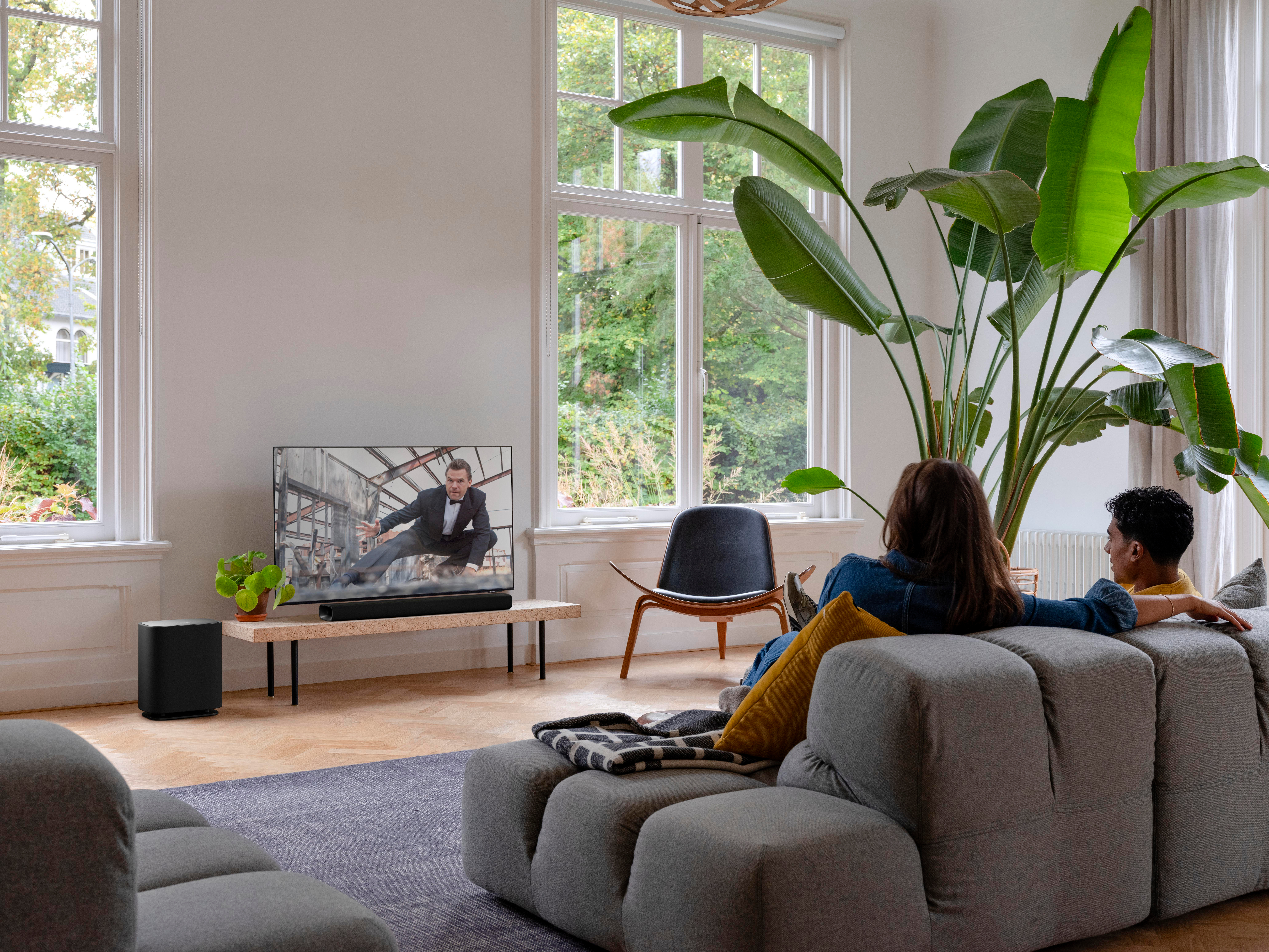 Ein Mann im Anzug auf einem Bildschirm, zwei Personen auf einem Sofa, ein schwarzer Subwoofer und Pflanzen.
