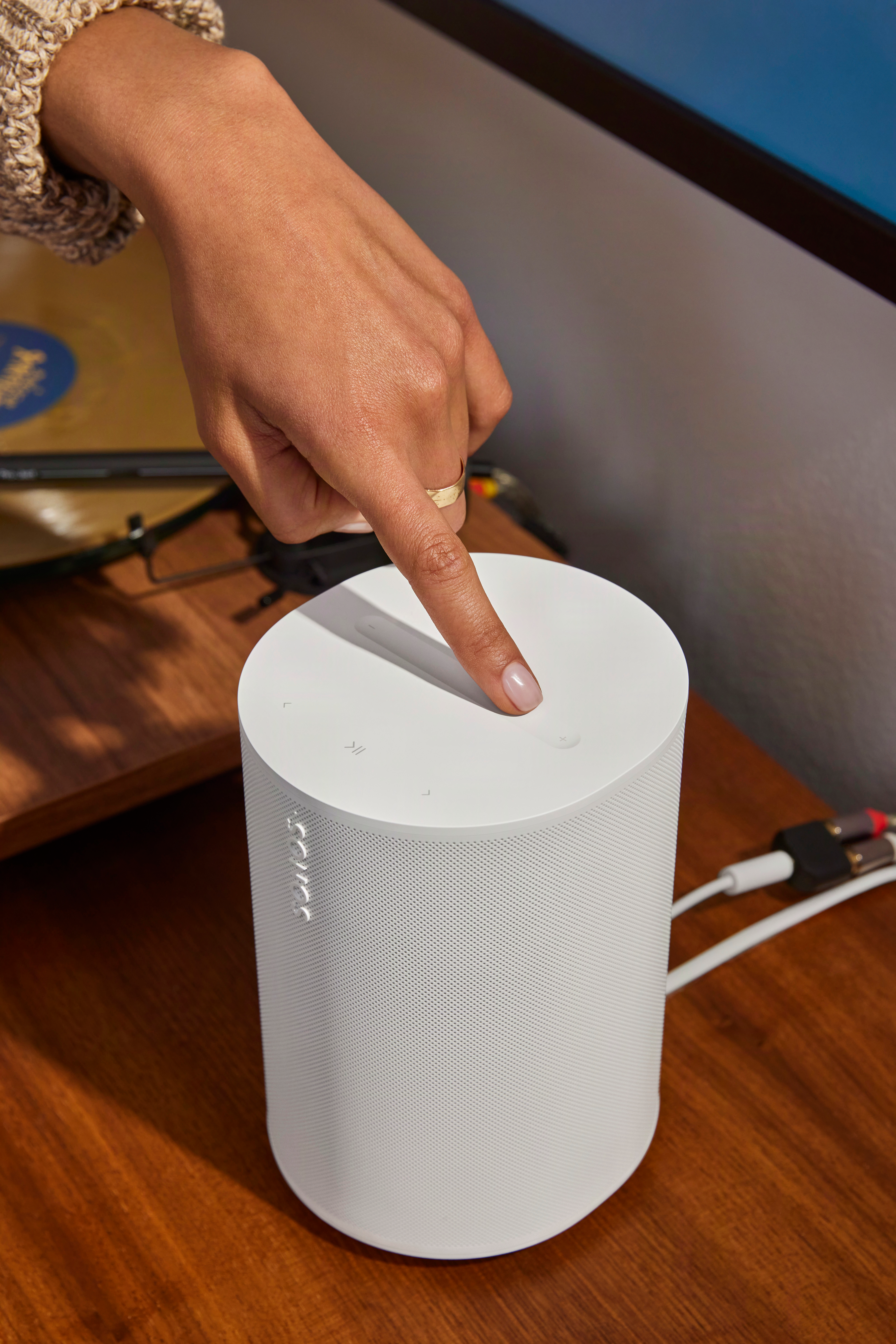 Eine Hand drückt auf einen weißen Sonos Lautsprecher auf einem Holztisch.