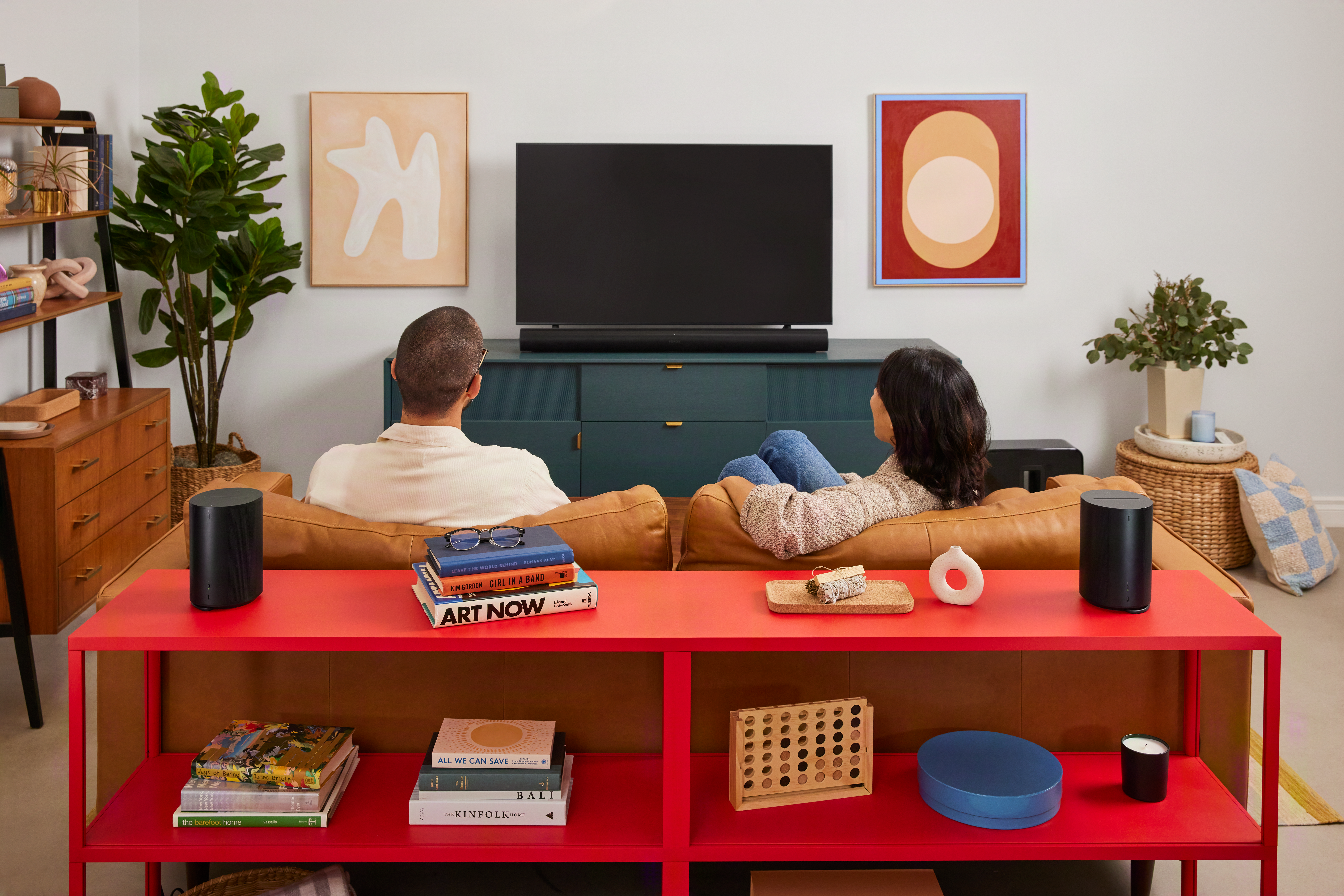 Paar auf Couch schaut Fernseher. Schwarze Lautsprecher, Bücher, Pflanzen, Kunst.