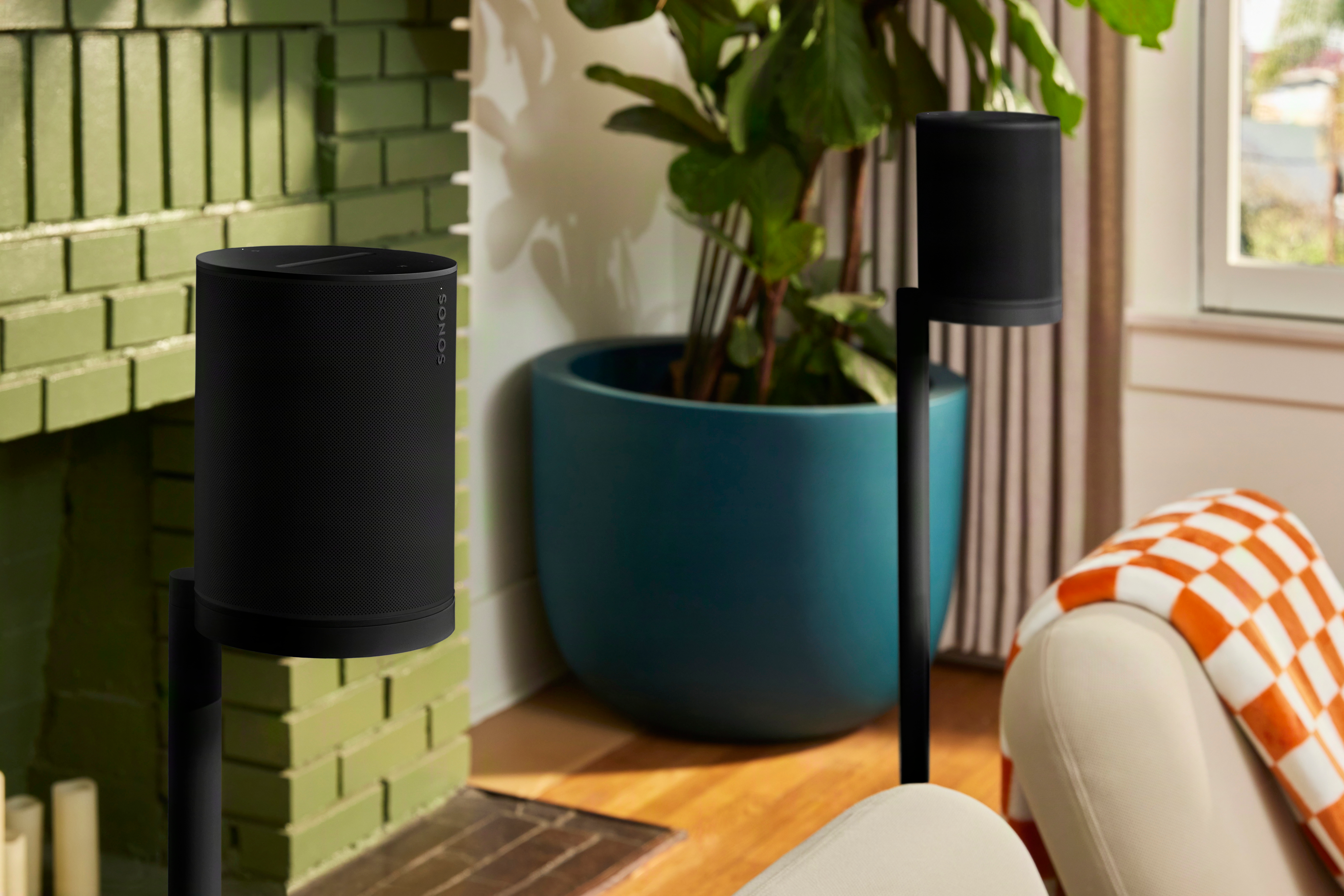 Zwei schwarze Sonos-Lautsprecher stehen auf Stativen vor einem grünen Kamin.