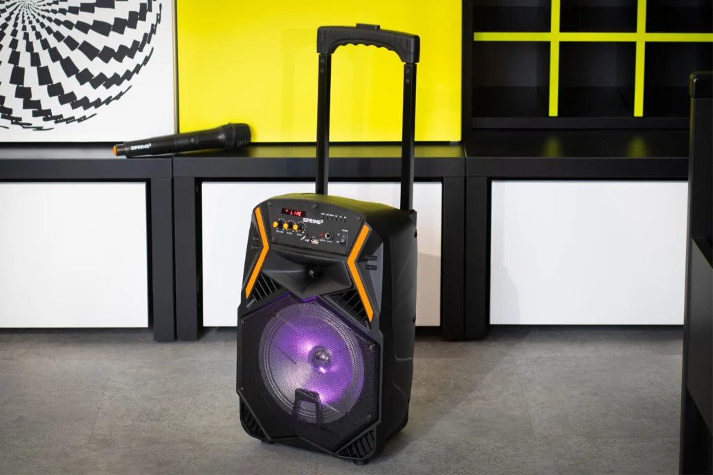 Przenośny głośnik z kółkami i mikrofonem na półce.