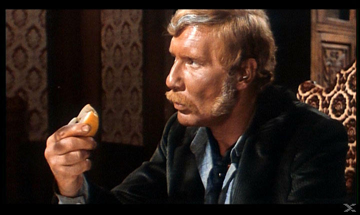 Ein Mann mit Schnurrbart hält ein Stück Orange in der Hand.