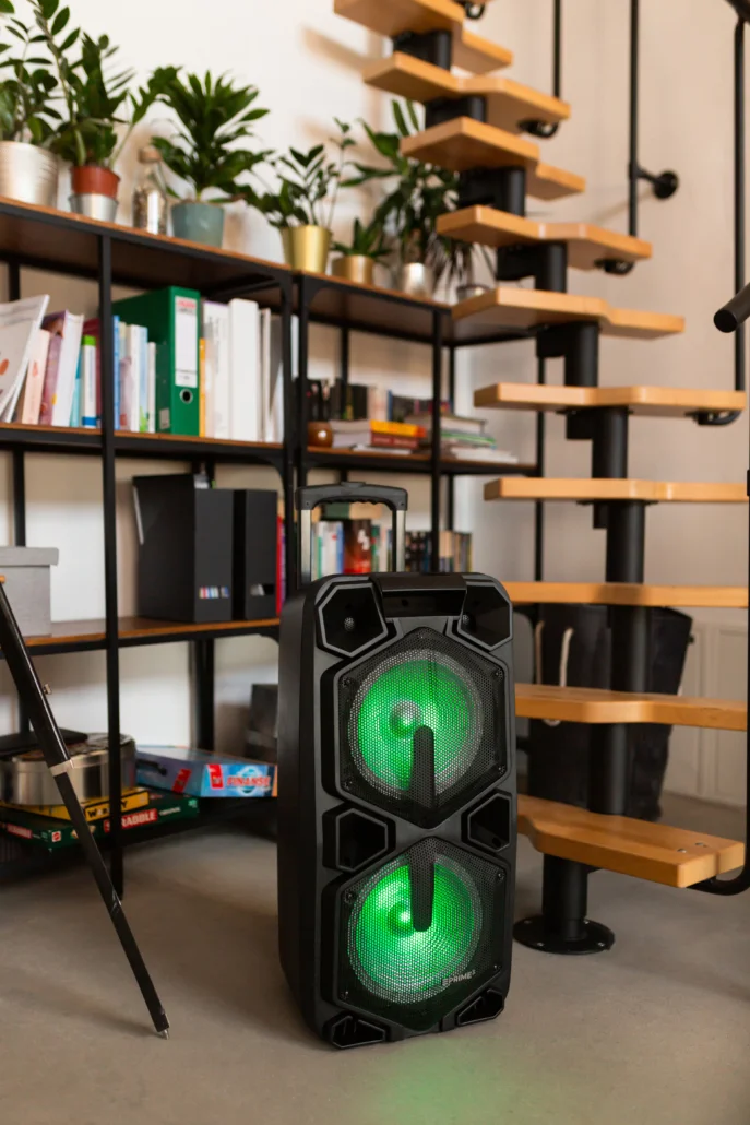 Czarny głośnik z zielonym podświetleniem stoi obok półki z książkami i roślinami.