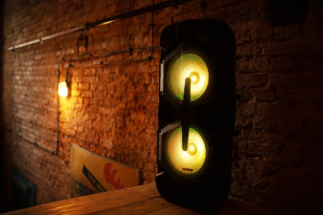 Czarny głośnik z dwoma jasnożółtymi przetwornikami stoi na drewnianej powierzchni przy ceglanym murze.