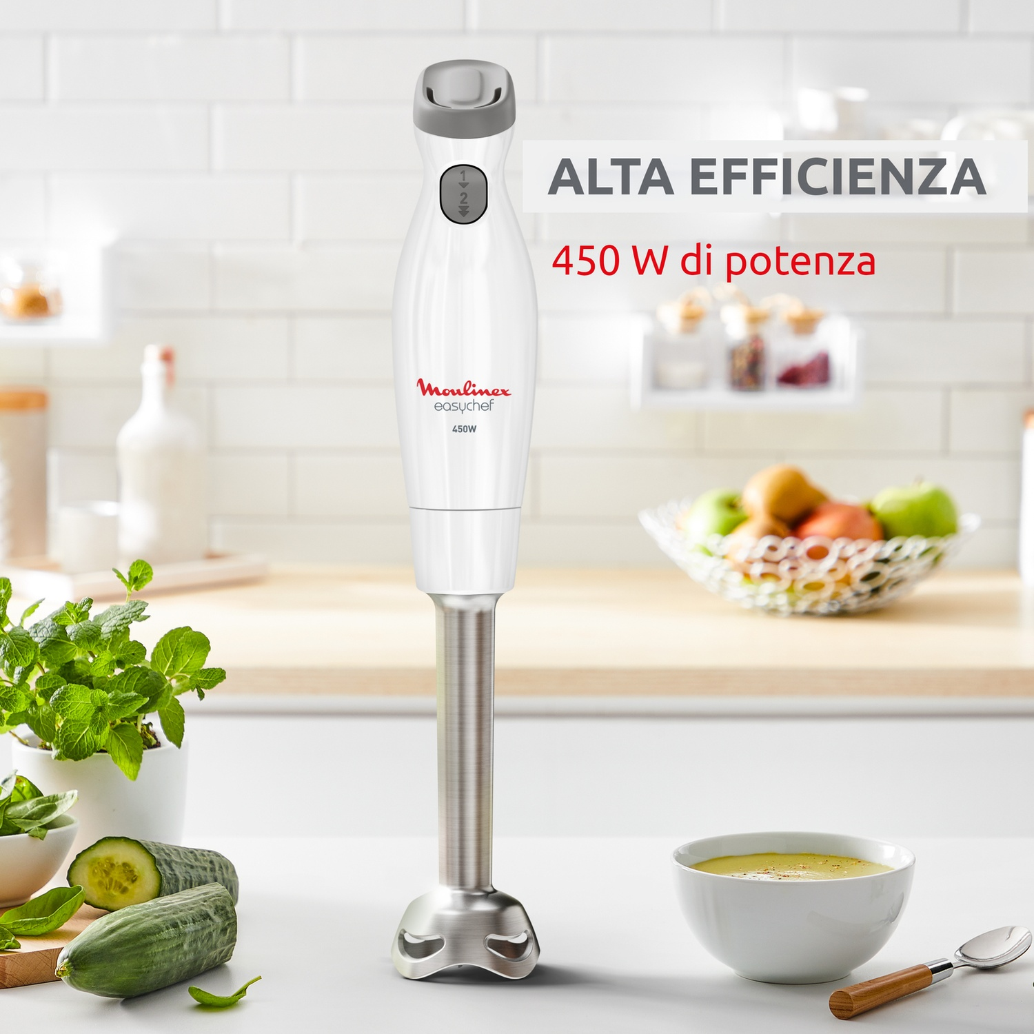 Un frullatore a immersione bianco poggia su un bancone con cetrioli, menta e una ciotola di zuppa.
