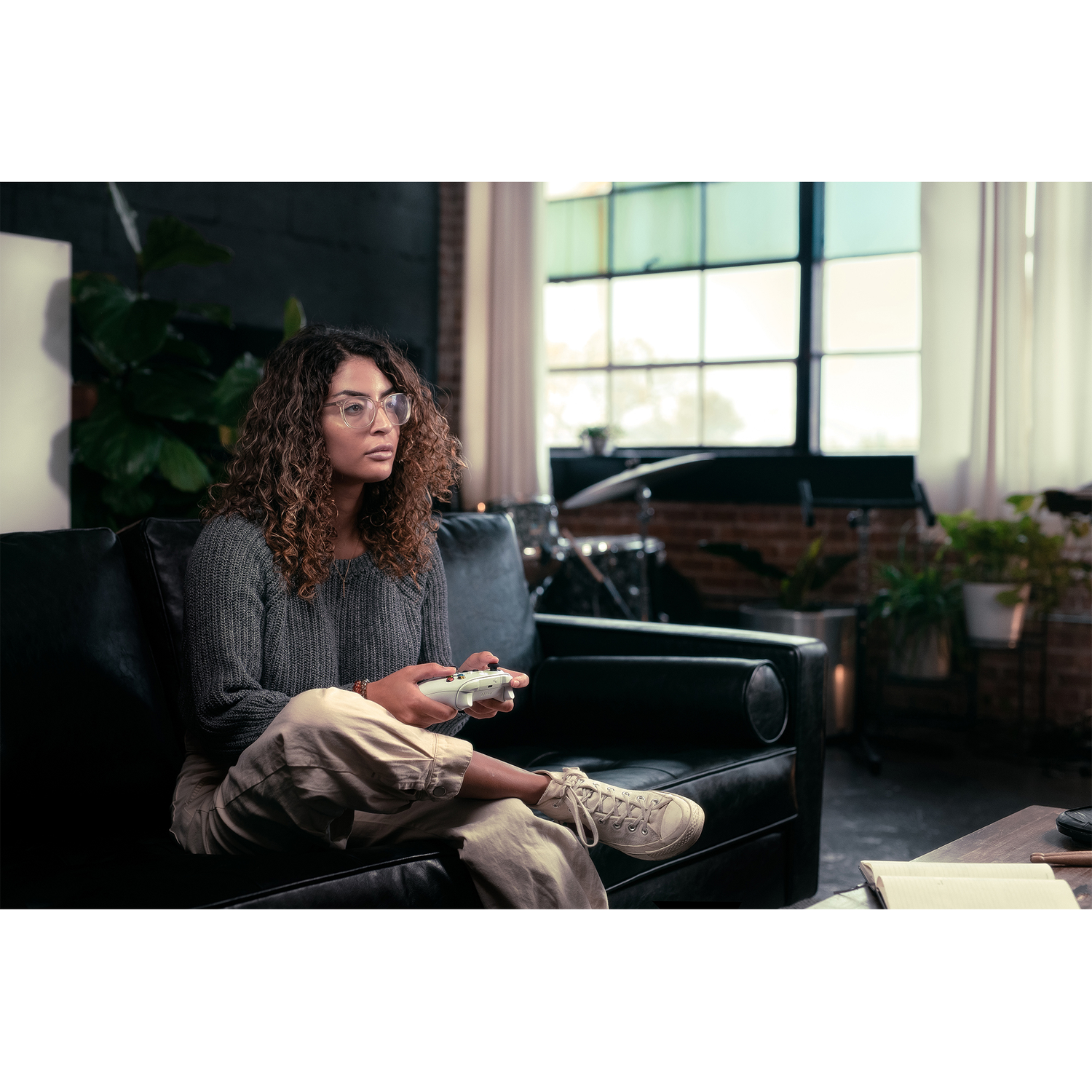 A woman with curly hair and glasses sits on a black couch, playing video games.