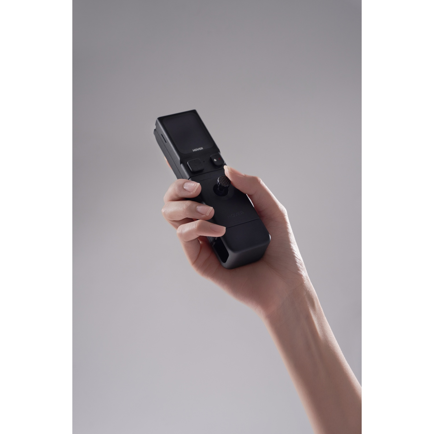A hand holds a black remote control with a screen and a joystick.