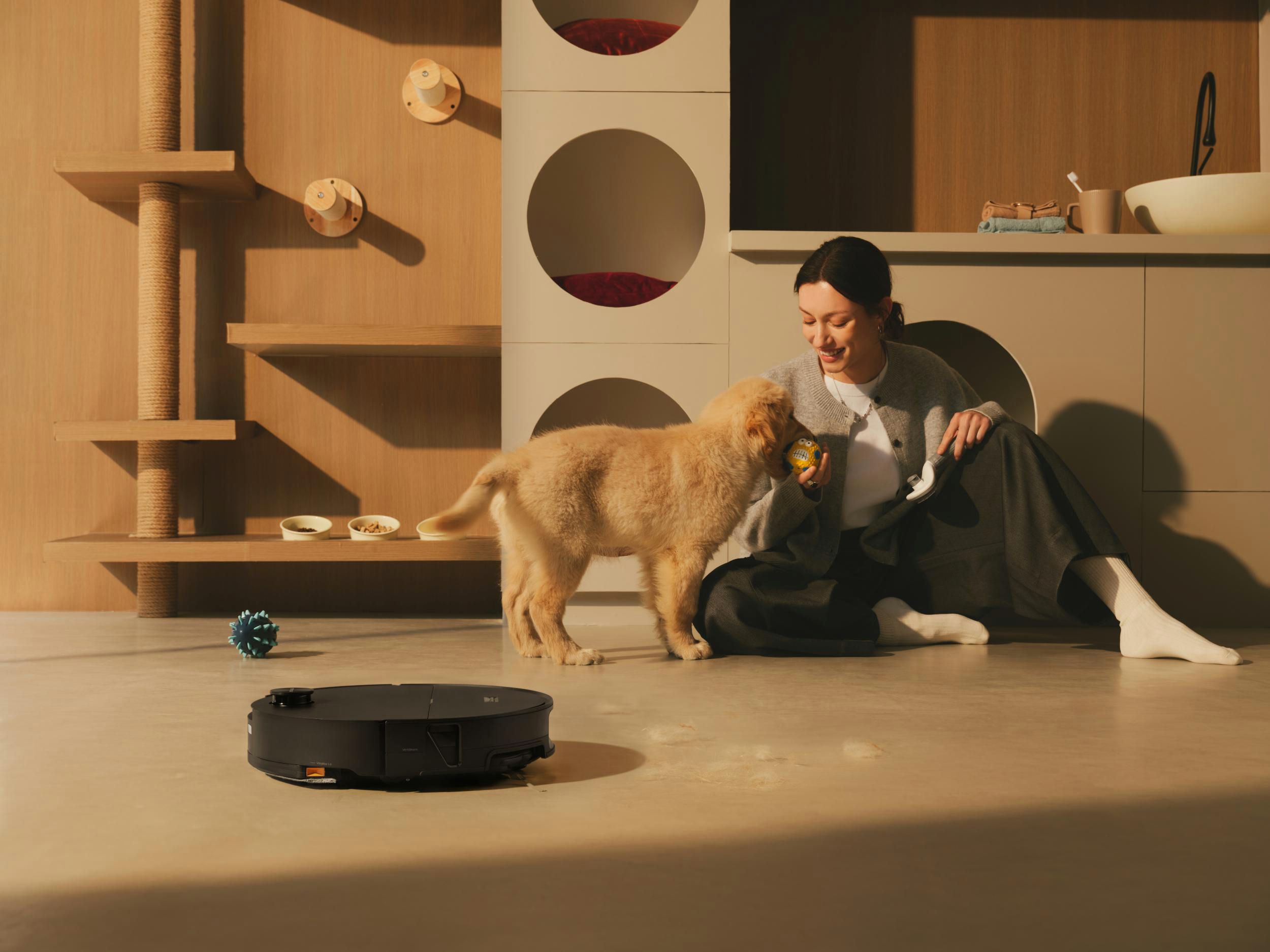 Ein Hund spielt mit einem Spielzeug, während eine Frau zusieht. Ein Saugroboter ist auf dem Boden.