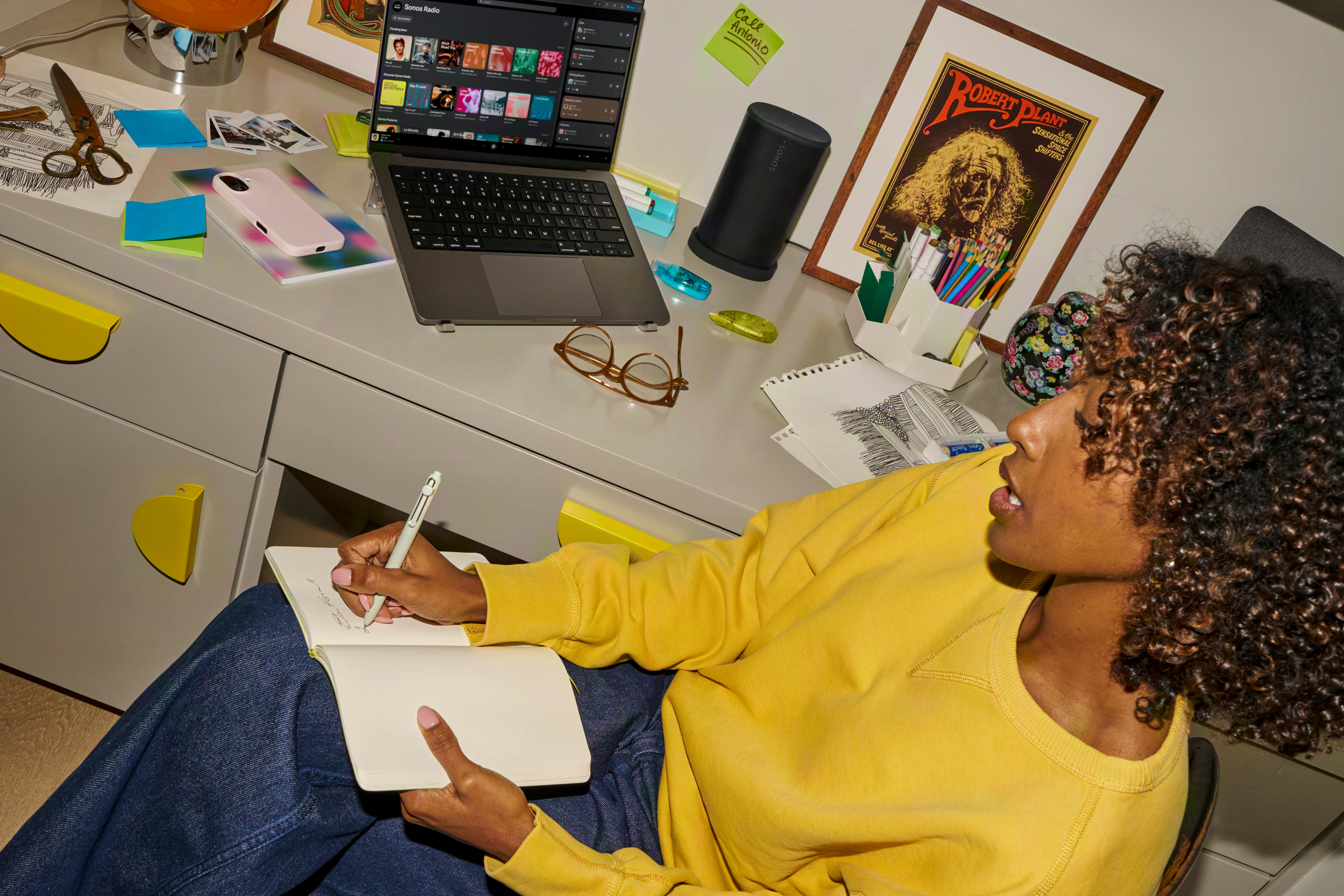 Une femme aux cheveux bouclés écrit dans un carnet. Un ordinateur portable et une enceinte sont sur le bureau.