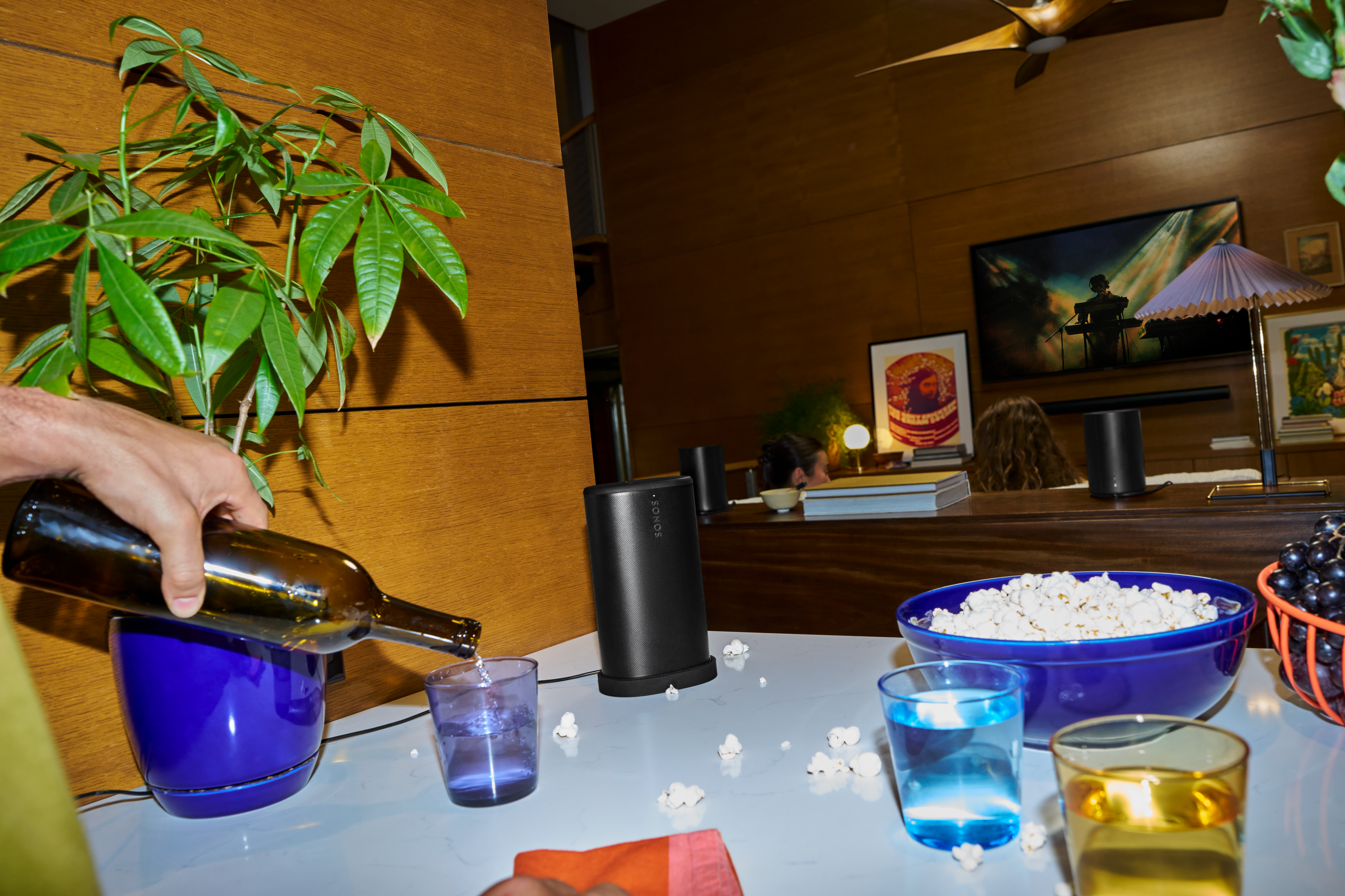 Une main verse d'une bouteille dans un verre. Popcorn, raisins et haut-parleurs sur la table.