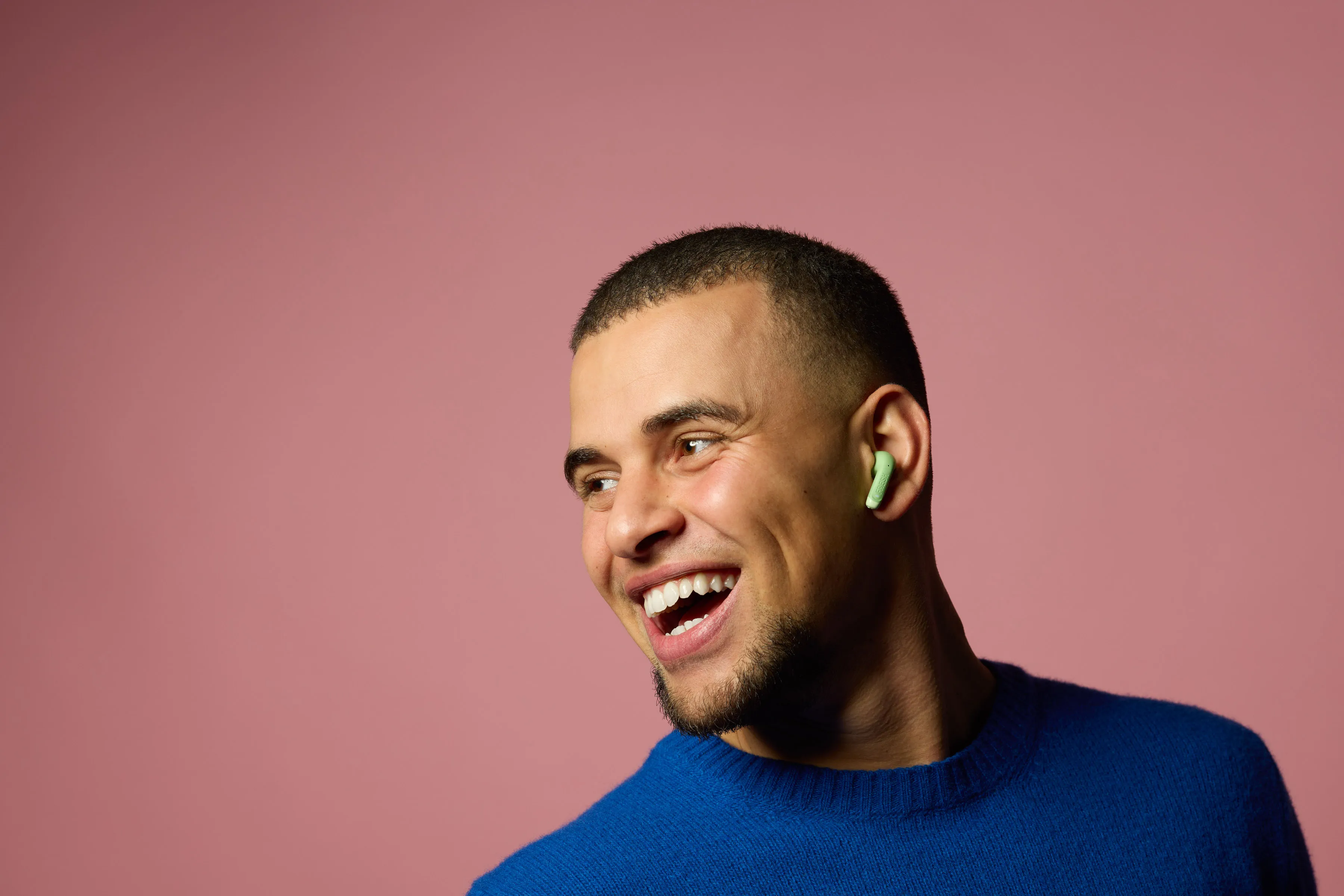 Ein Mann in einem blauen Pullover lacht mit einem grünen Ohrhörer im Ohr.