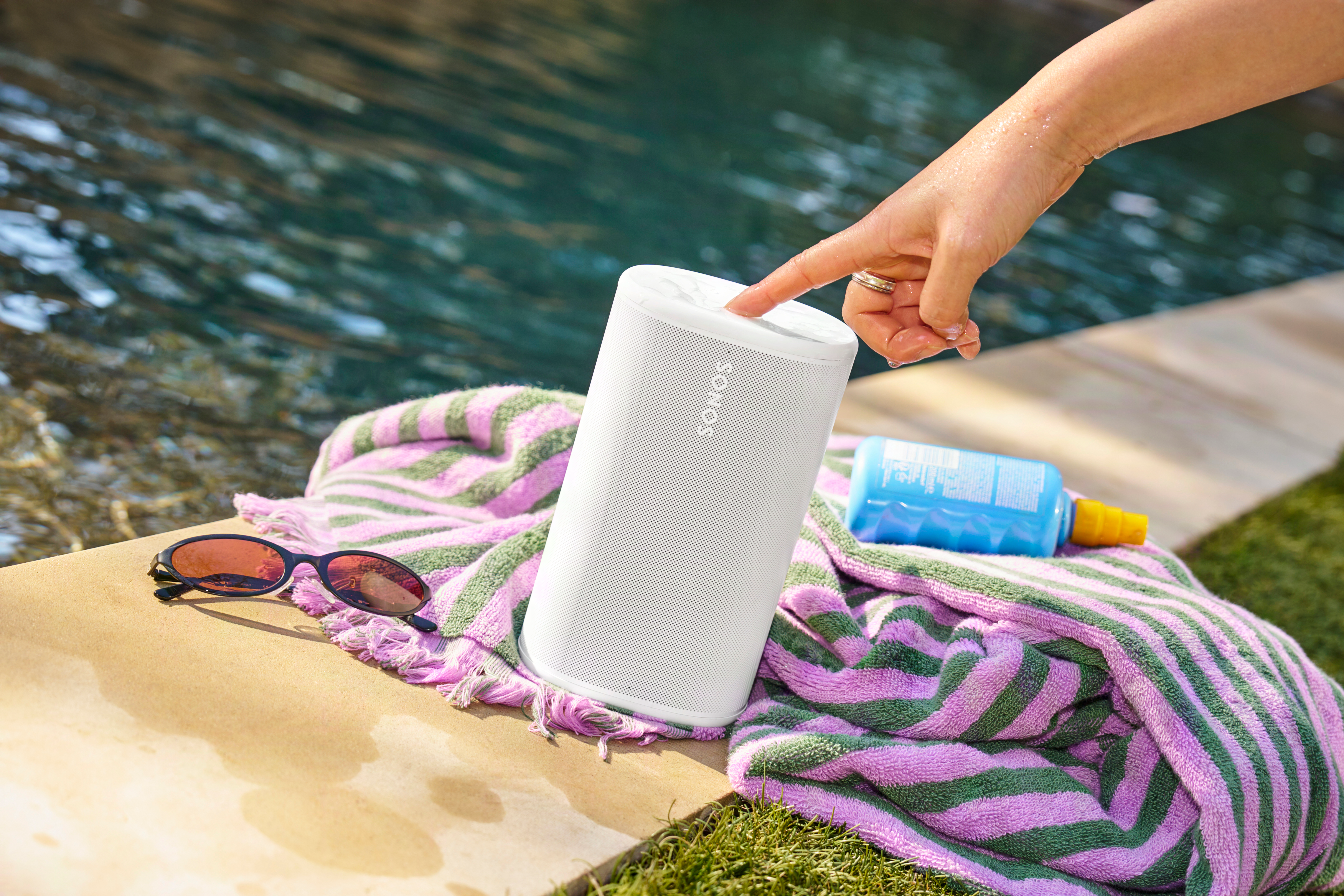 Une main tape sur une enceinte blanche près d'une piscine. Lunettes de soleil, serviette et crème solaire à proximité.