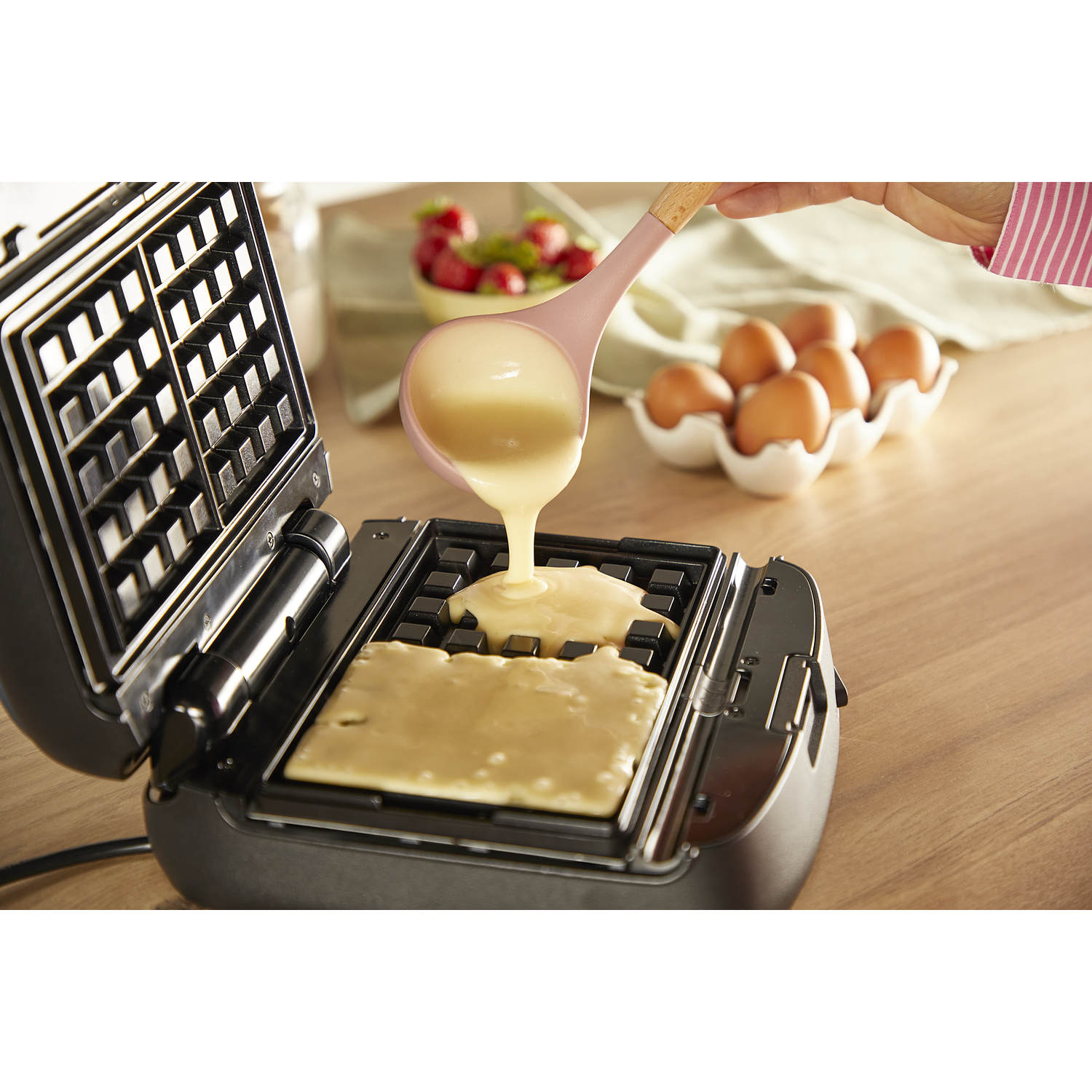 Batter being poured into an open waffle maker. Eggs and strawberries in background.