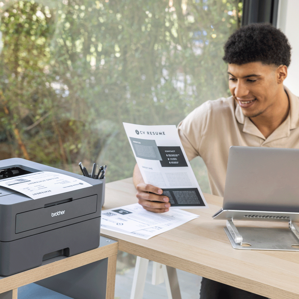 Mann liest Lebenslauf neben Brother-Drucker und Laptop im Büro.