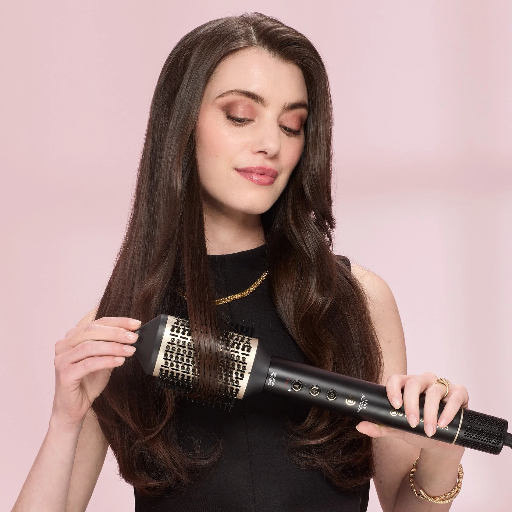 Una mujer con cabello oscuro se peina con un cepillo caliente negro y dorado sobre fondo rosa.