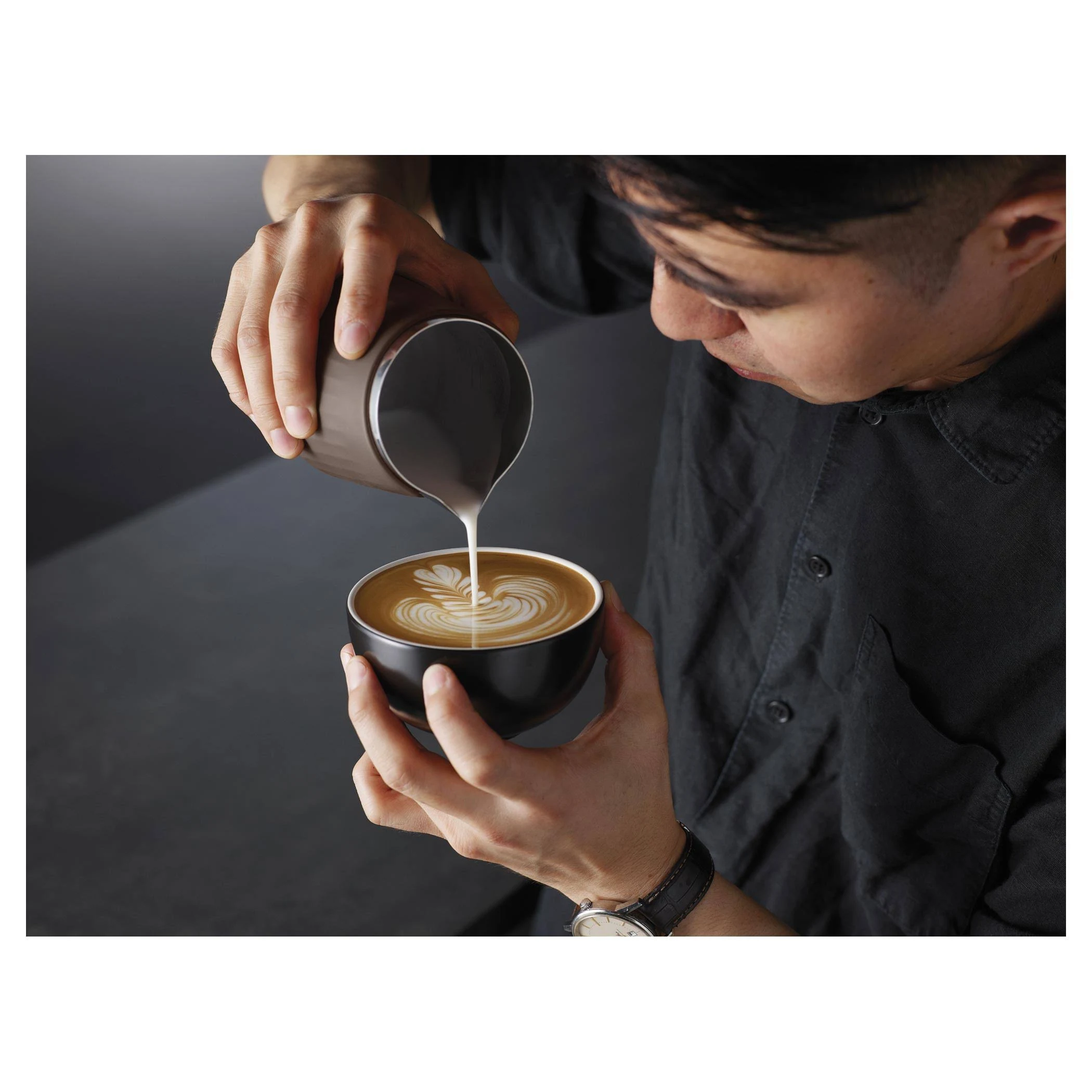 Barista vierte arte latte en un tazón negro, creando un patrón de hoja con espuma blanca.
