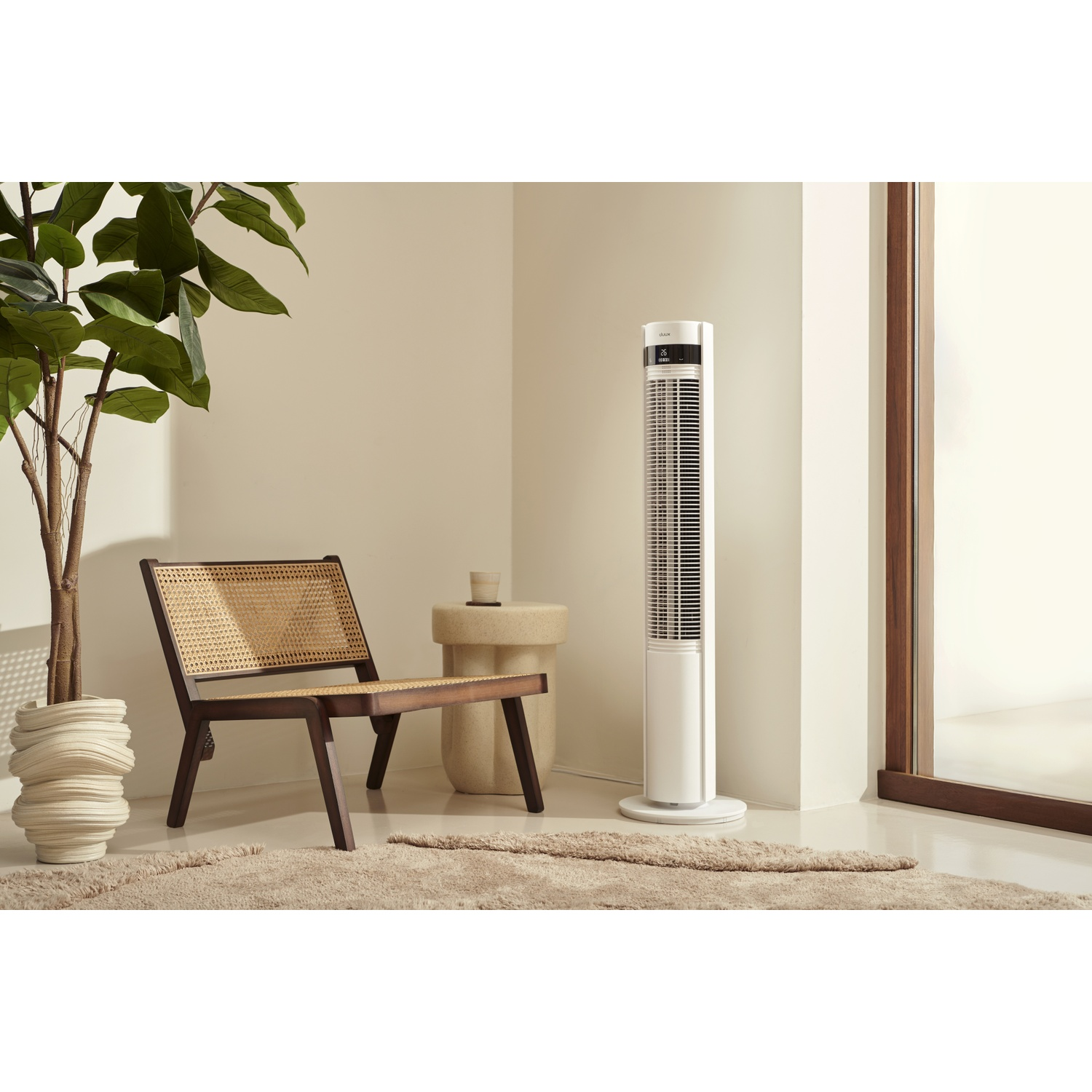 A modern white tower fan stands next to a cane chair and a large plant.