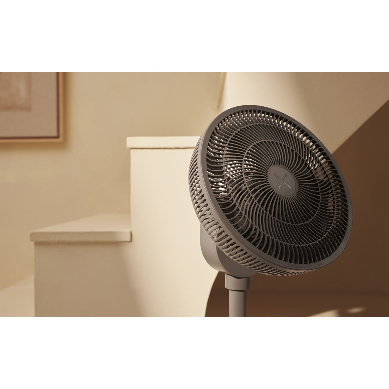 Un ventilateur sur pied gris est placé à côté d'un escalier beige clair avec un tableau.