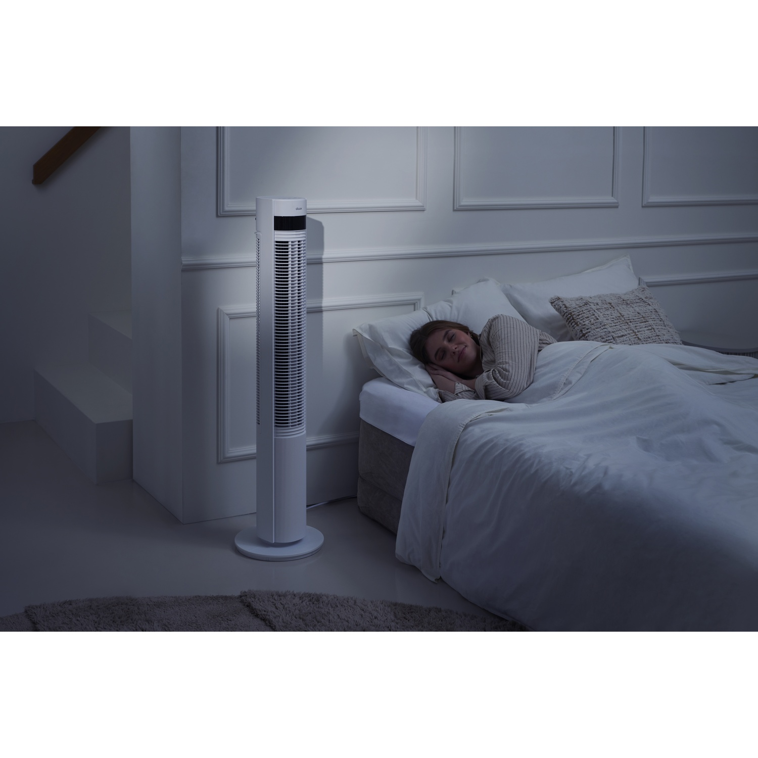 Une femme dort dans un lit, un ventilateur sur pied à côté.