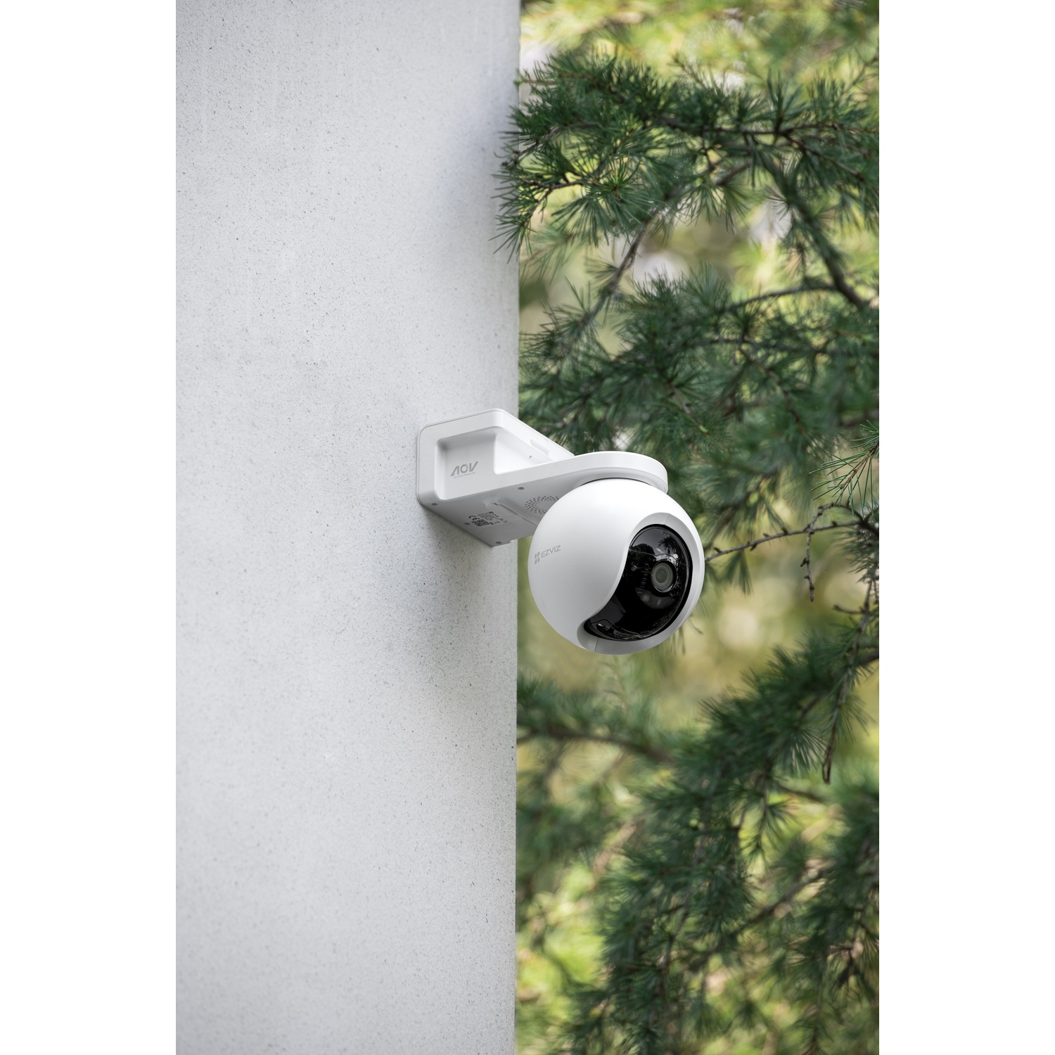 A white security camera is mounted on a gray wall, with trees in the background.
