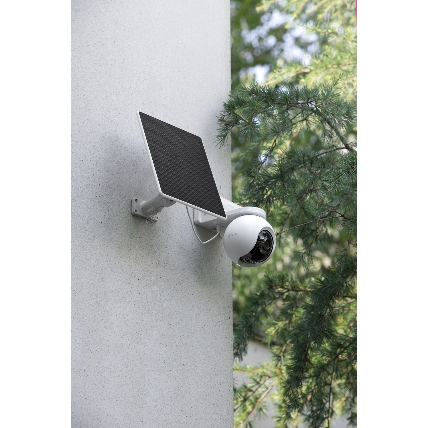 White security camera with solar panel mounted on a wall, next to green trees.