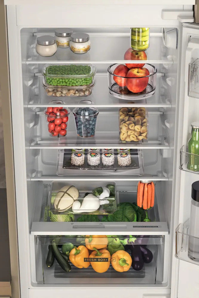 Otwarta lodówka z jedzeniem na wielu półkach. Górne półki z słoikami, groszkiem i jabłkami. Dolne z sushi, warzywami, paprykami.