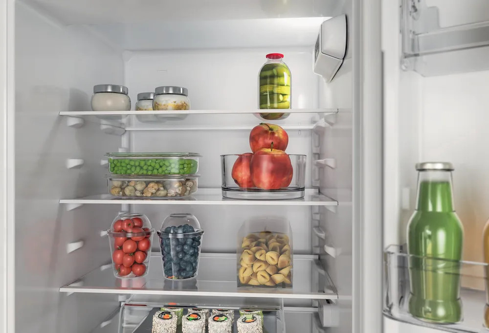 Otwarta lodówka z jedzeniem: słoiki, jabłka, groszek, pomidory, jagody, makaron i napoje.