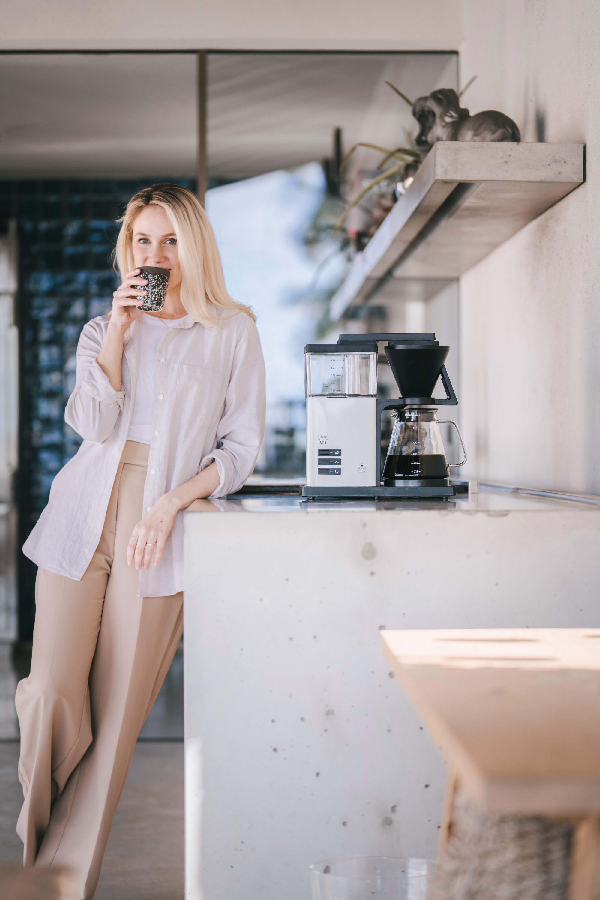 Una donna bionda beve da una tazza accanto a una macchina da caffè.