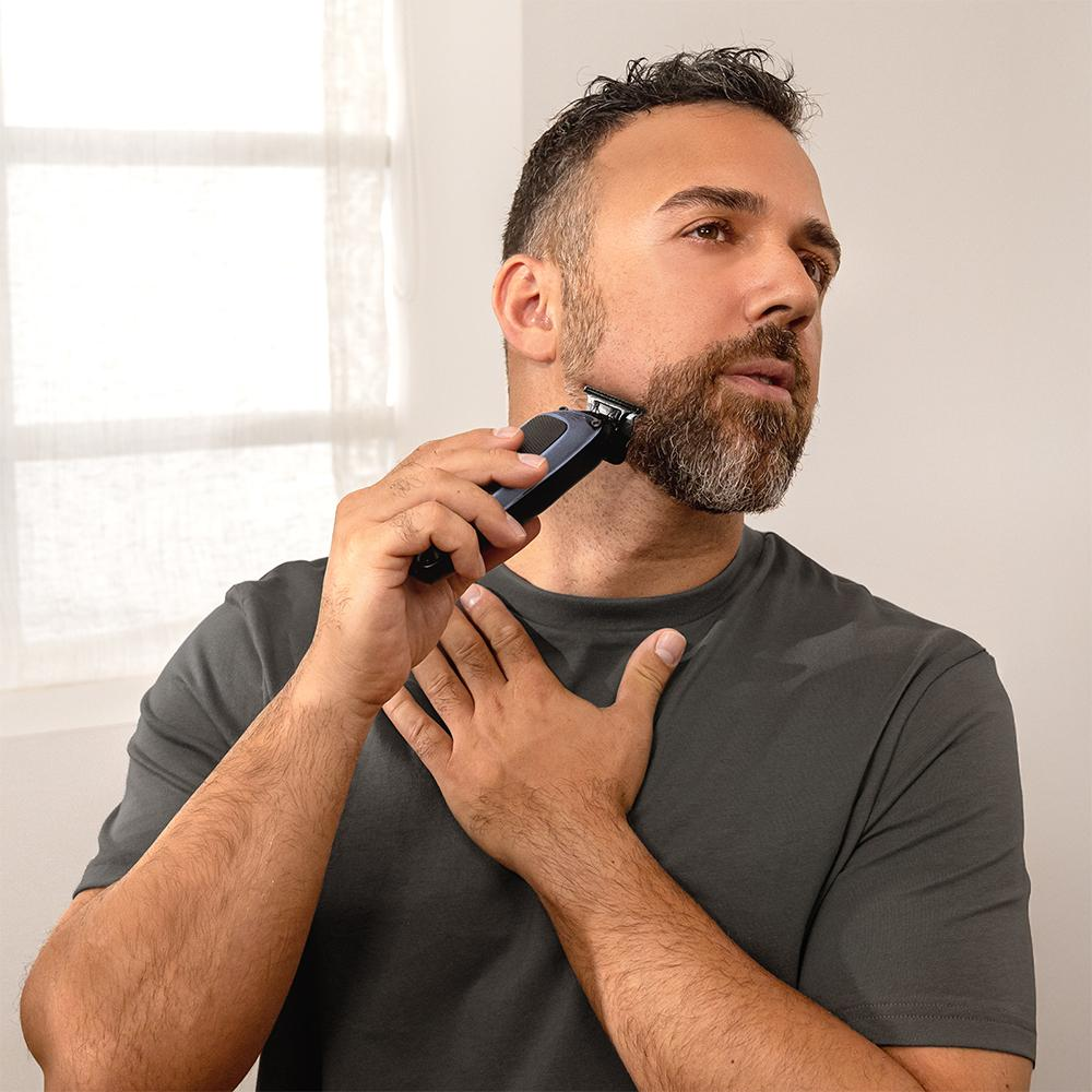 Un hombre se recorta la barba con una recortadora cerca de una ventana.