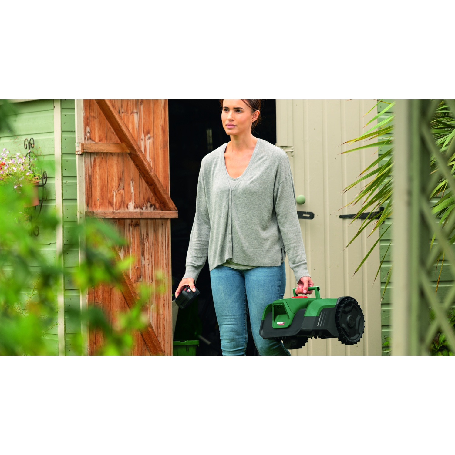Una donna porta un tosaerba verde e una batteria davanti a un capanno da giardino.