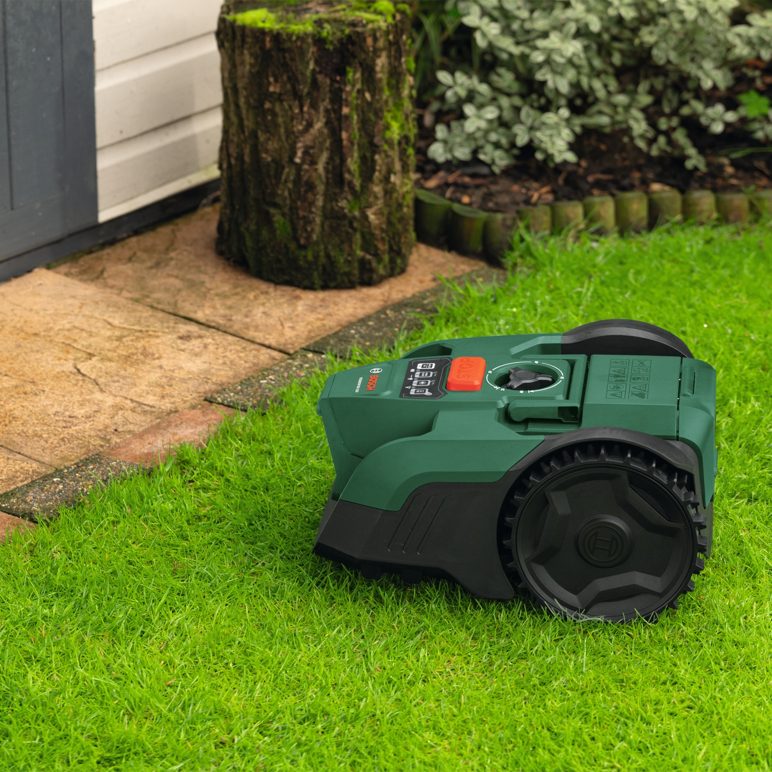 Un tosaerba robotizzato verde è sull'erba accanto a un ceppo e a un sentiero.