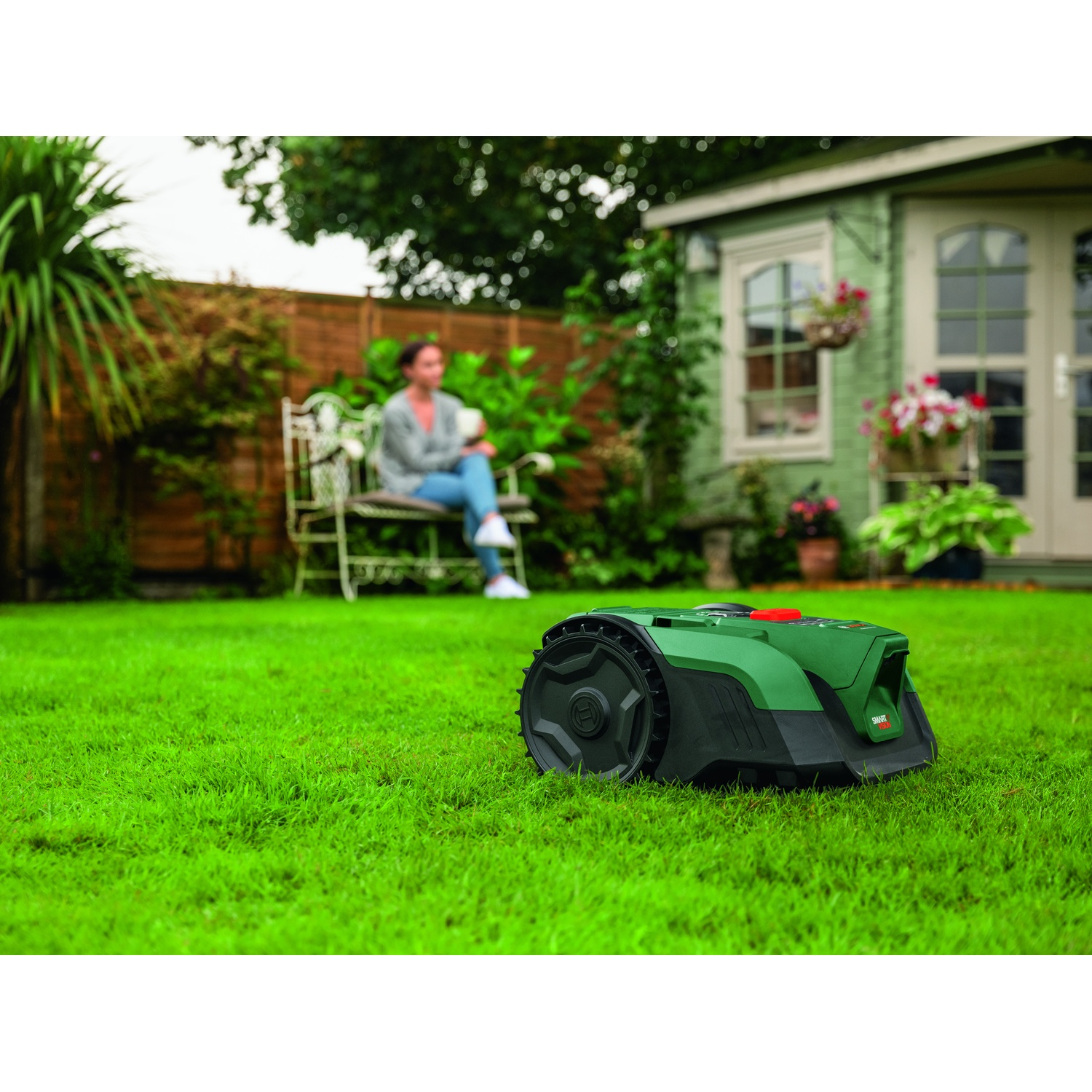 Un robot tosaerba verde è sull'erba. Una donna è seduta su una panchina sullo sfondo.