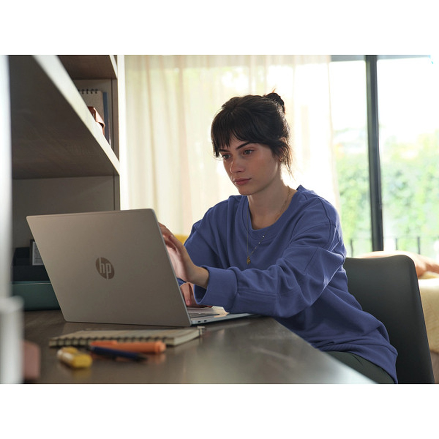 A young woman with her hair in a bun sits at a desk using a laptop computer.