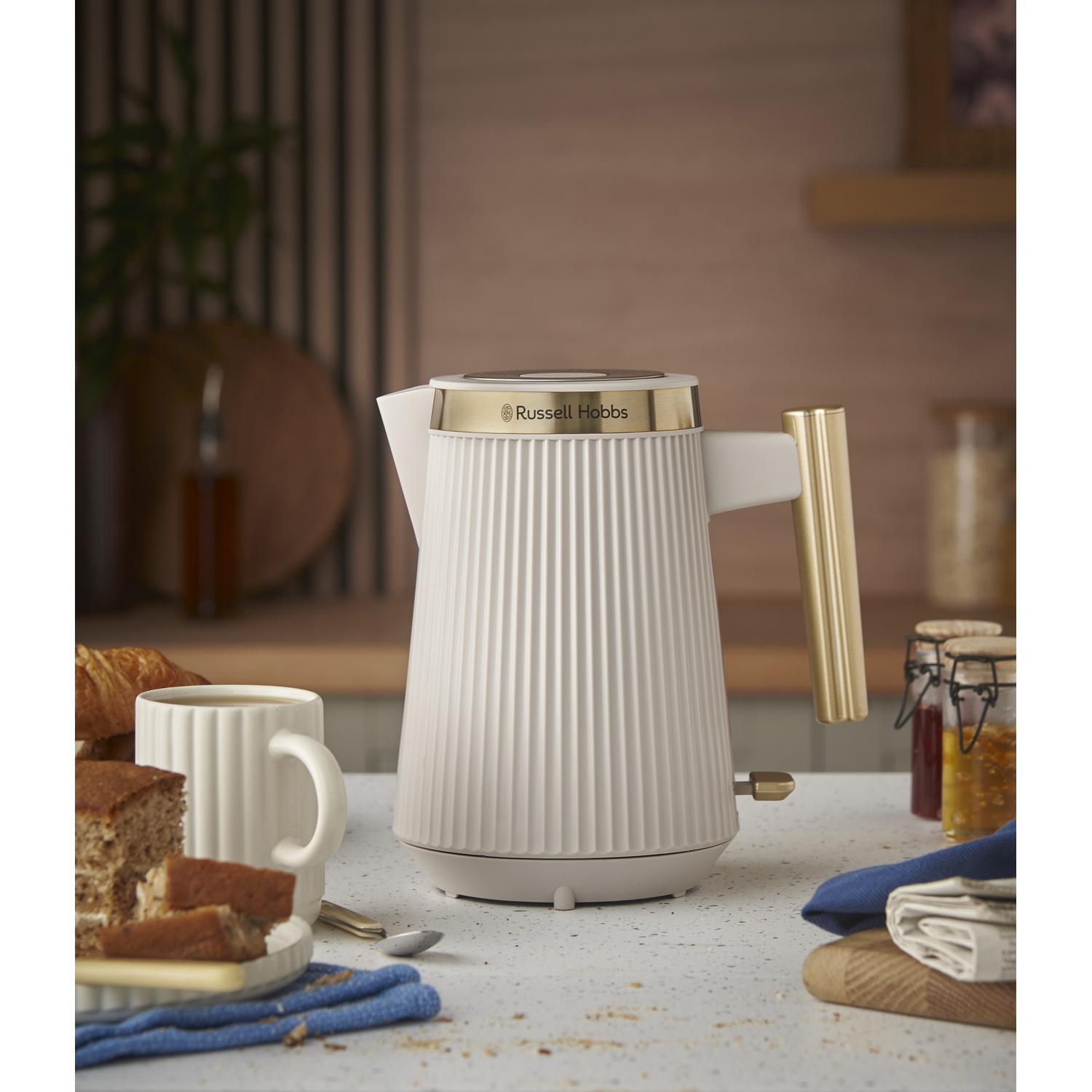 A white kettle with gold accents sits on a table with breakfast items.
