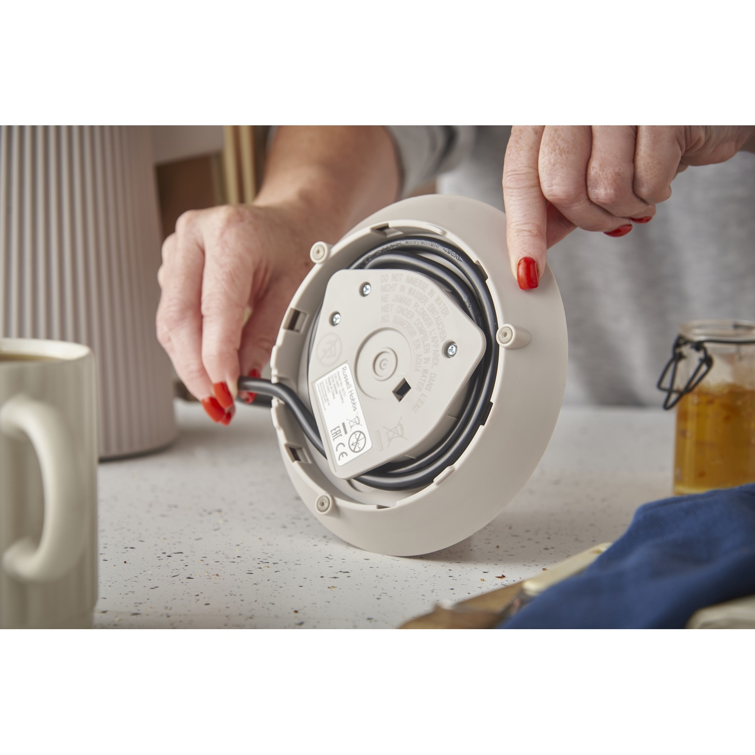 A hand with red nails holds the base of a kettle, with the cord visible.