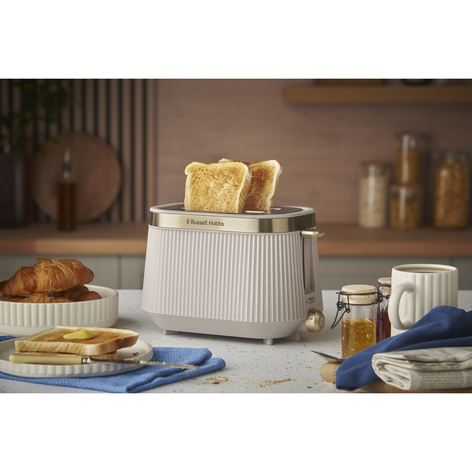 A beige toaster with two toast slices, croissants, jam, and coffee nearby.