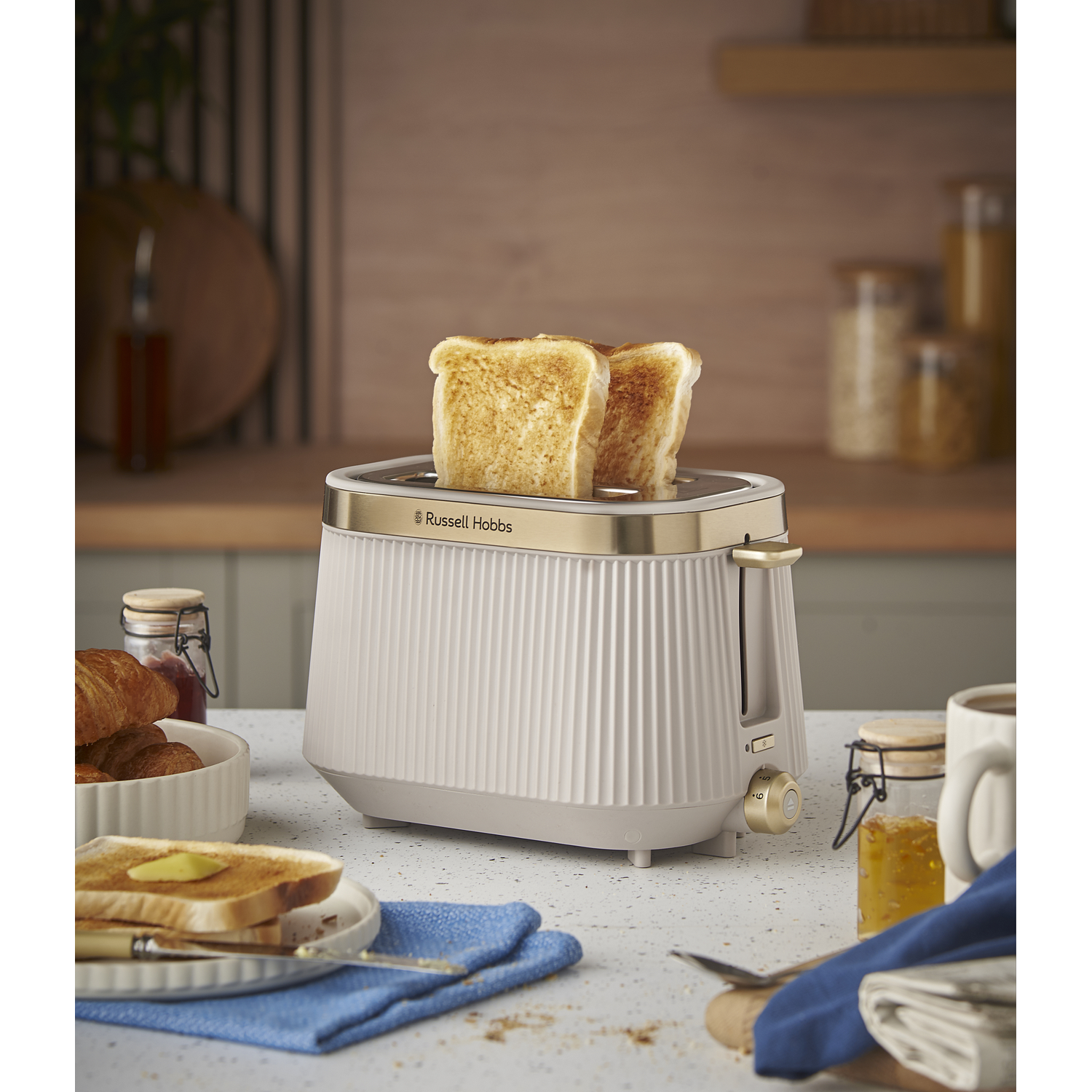 A cream-colored toaster with two slices of toast, croissants, and jam nearby.