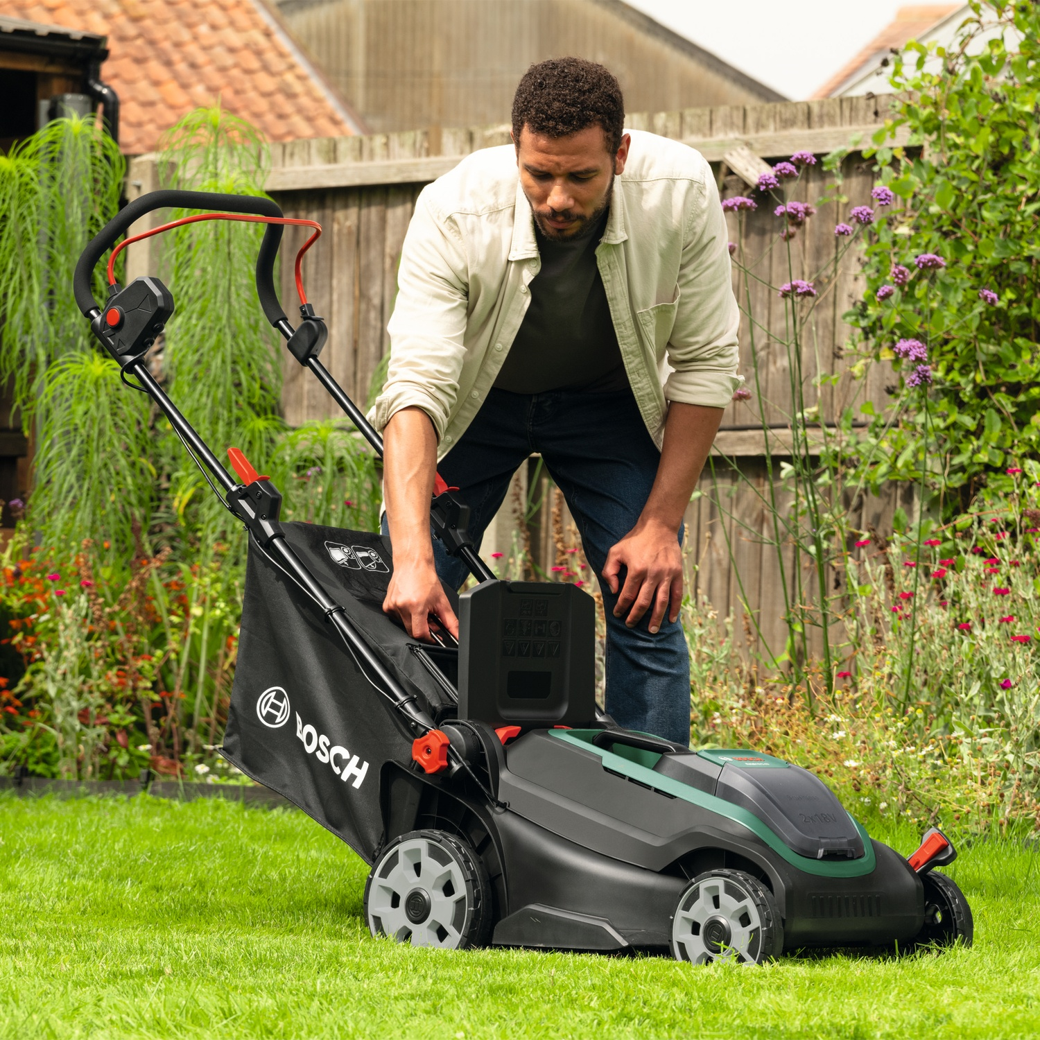 Uomo in giardino regola un tosaerba Bosch nero su erba verde con recinzione in legno dietro.