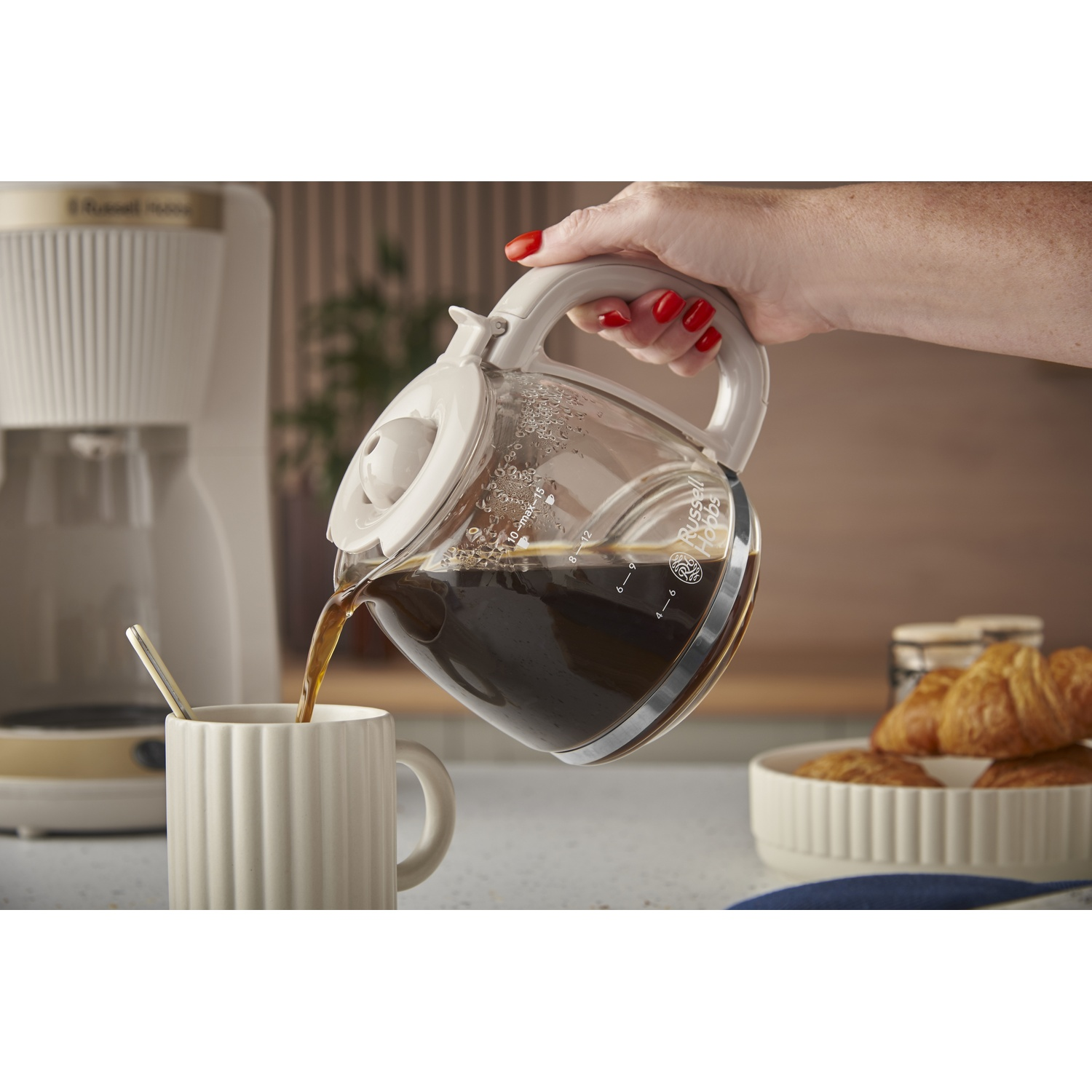 Een koffiepot schenkt koffie in een mok, met croissants en een koffiezetapparaat in de buurt.