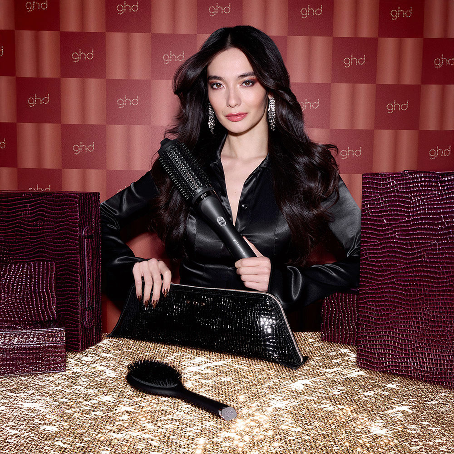 A woman with long dark hair holds a black curling iron on a sparkly surface.