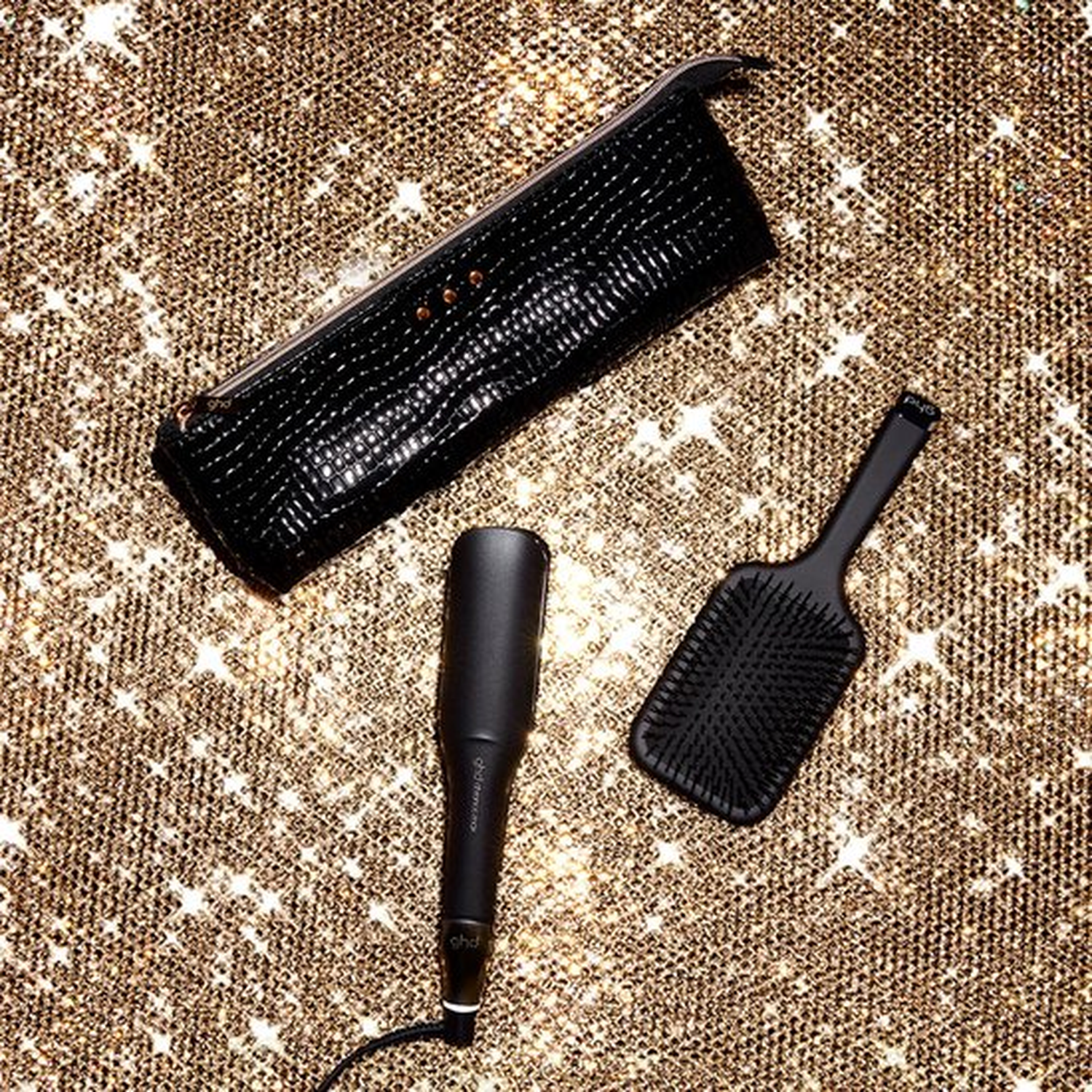 Black GHD hair straightener, brush, and case on a sparkly gold background.